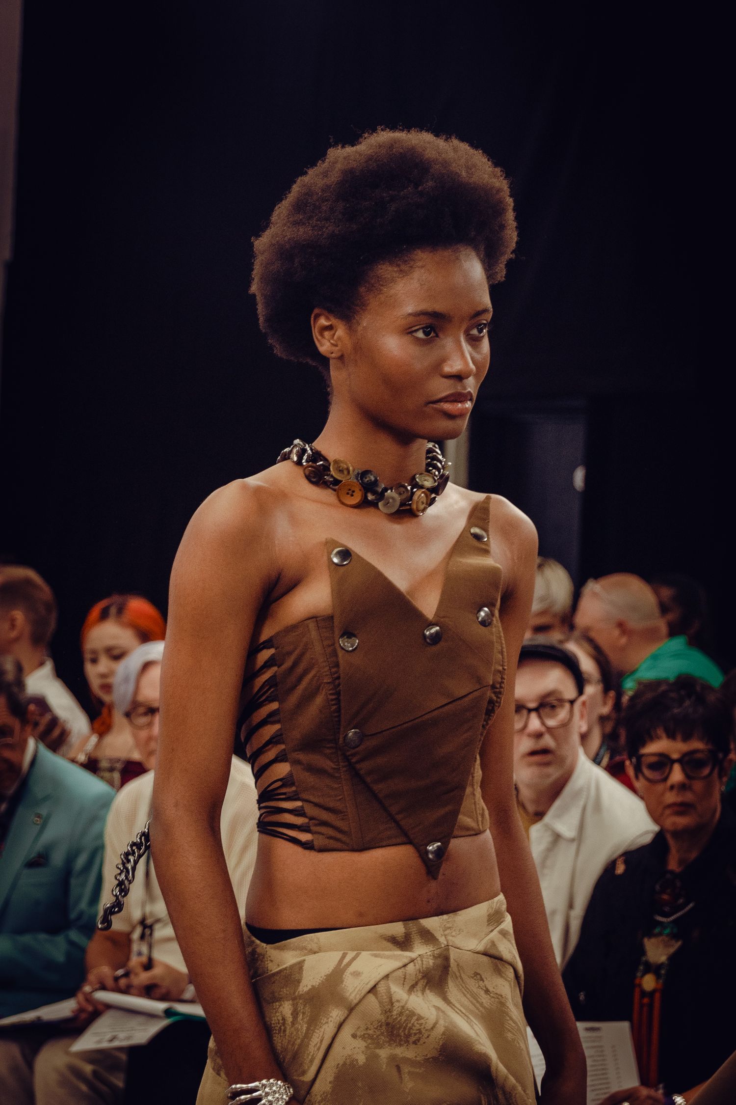 Model presents brown studded corset top with statement necklace at fashion show.