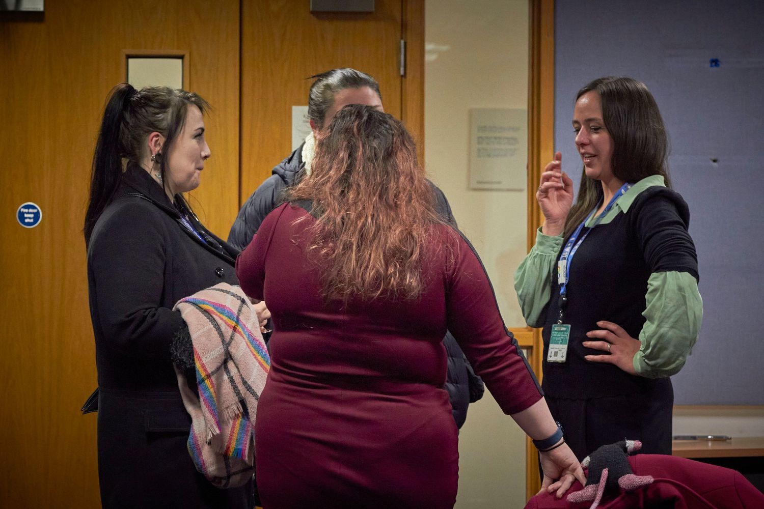 Three people having an animated conversation in an indoor hallway near a closed door.