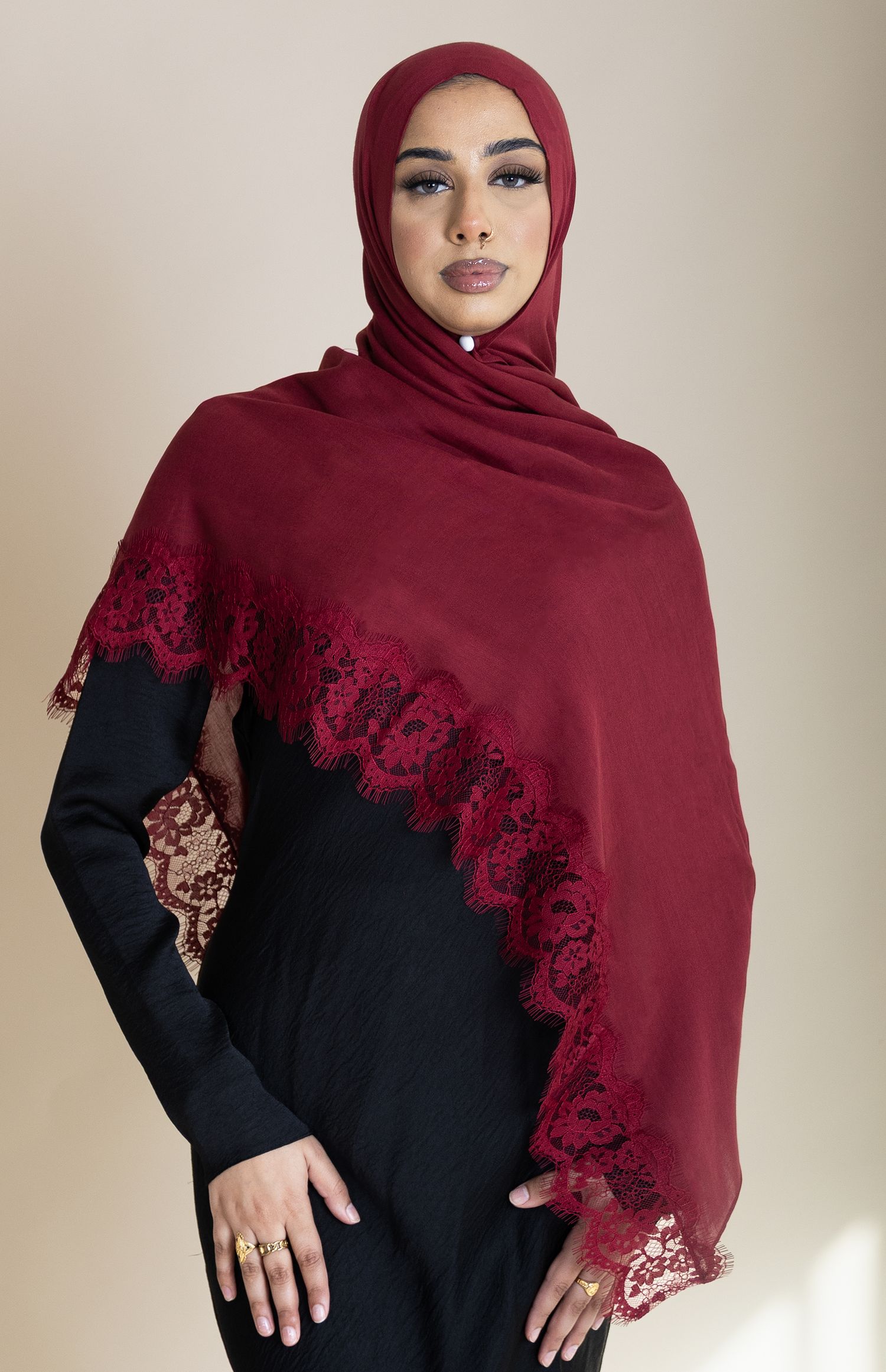 Woman wearing a burgundy lace-trimmed hijab and black dress poses elegantly against a neutral background.