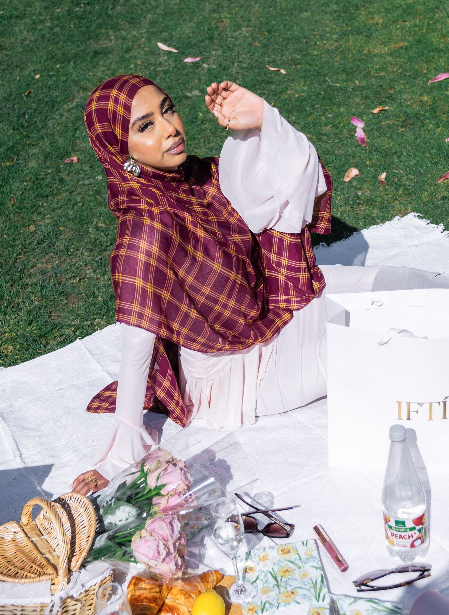Woman in burgundy plaid hijab and white outfit sitting on a picnic blanket outdoors with flowers and water bottle.