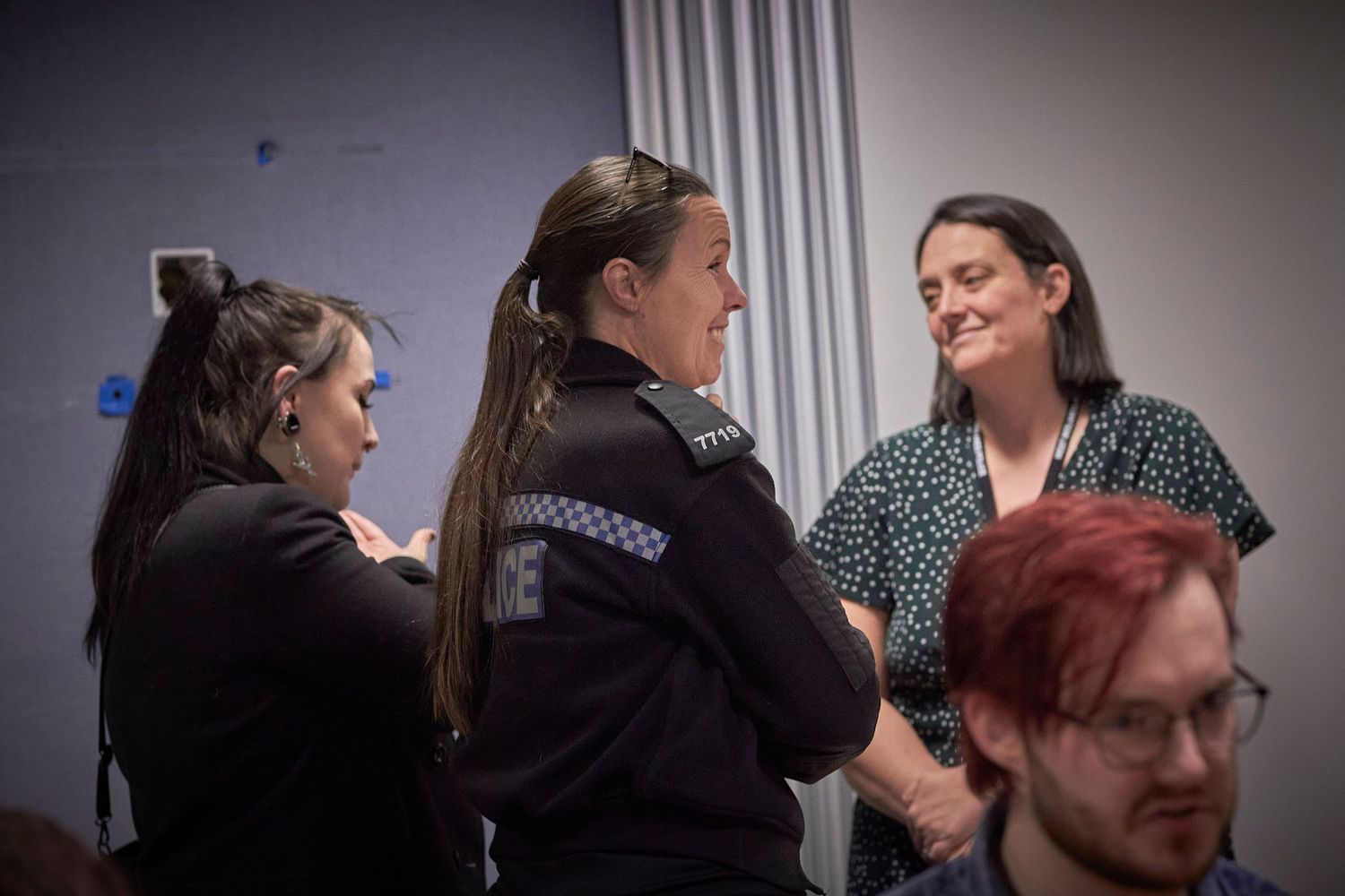 Three people in conversation, with a police officer in uniform visible, during an indoor meeting.