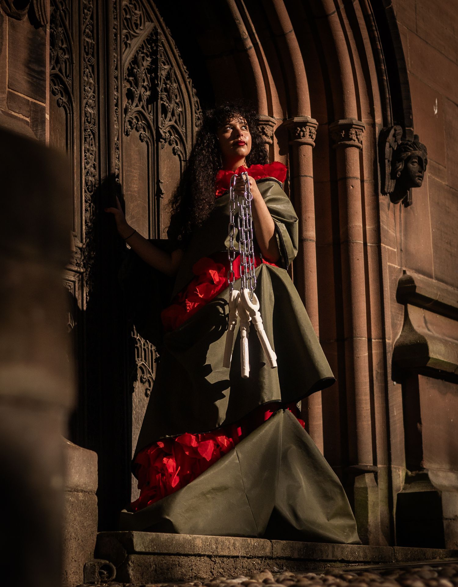 Woman in green gown holding red roses stands dramatically at gothic stone church entrance in dark moody lighting.