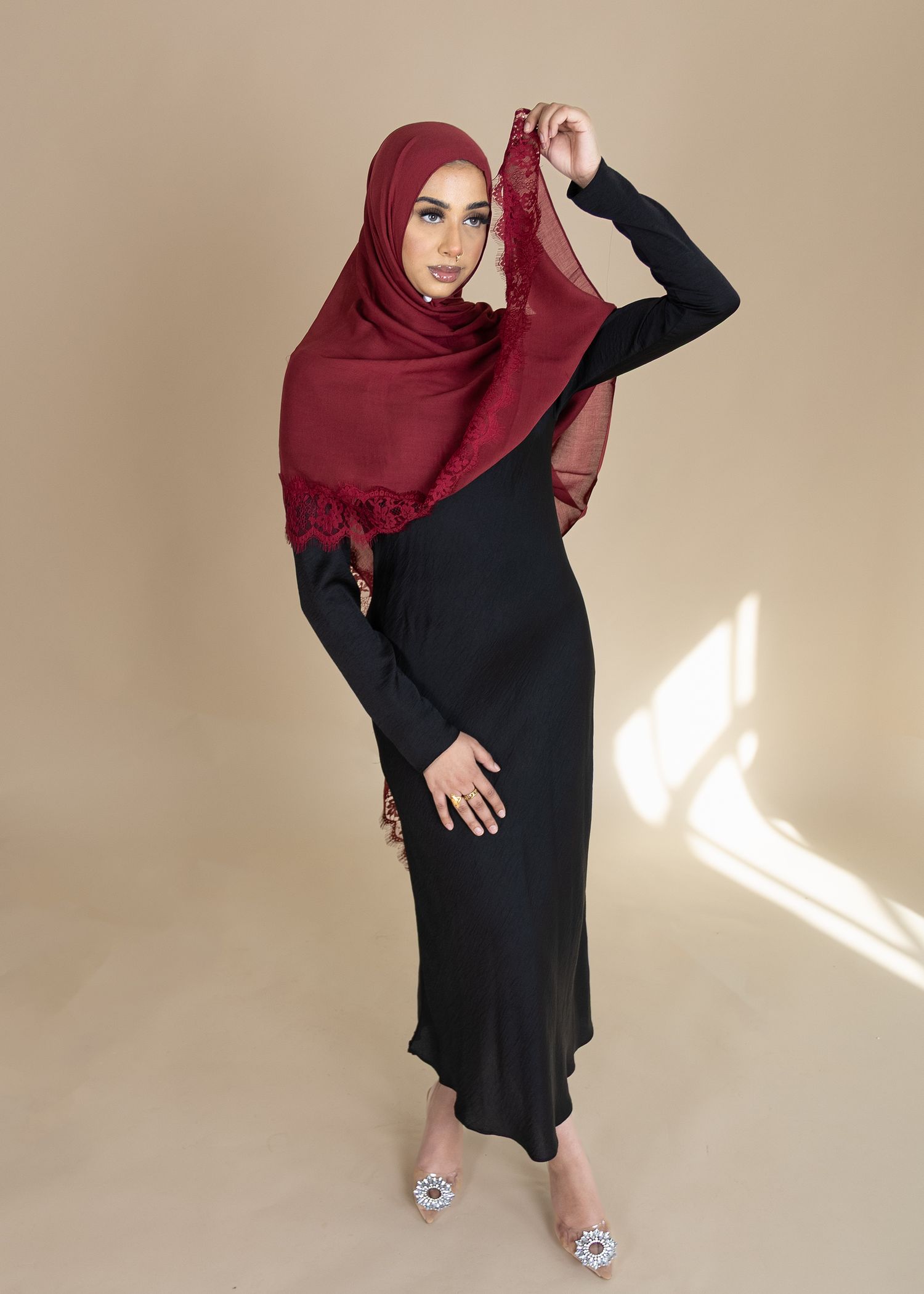 A woman in a burgundy hijab and black dress poses in different angles against a beige wall.