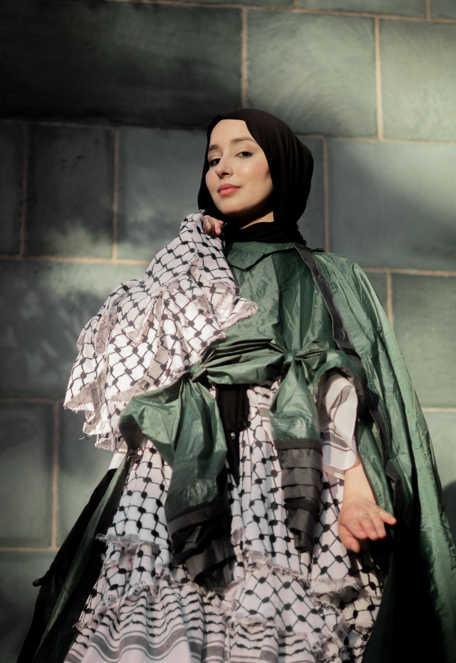 Fashion portrait featuring green and patterned keffiyeh-style garments against a stone wall in dramatic lighting.