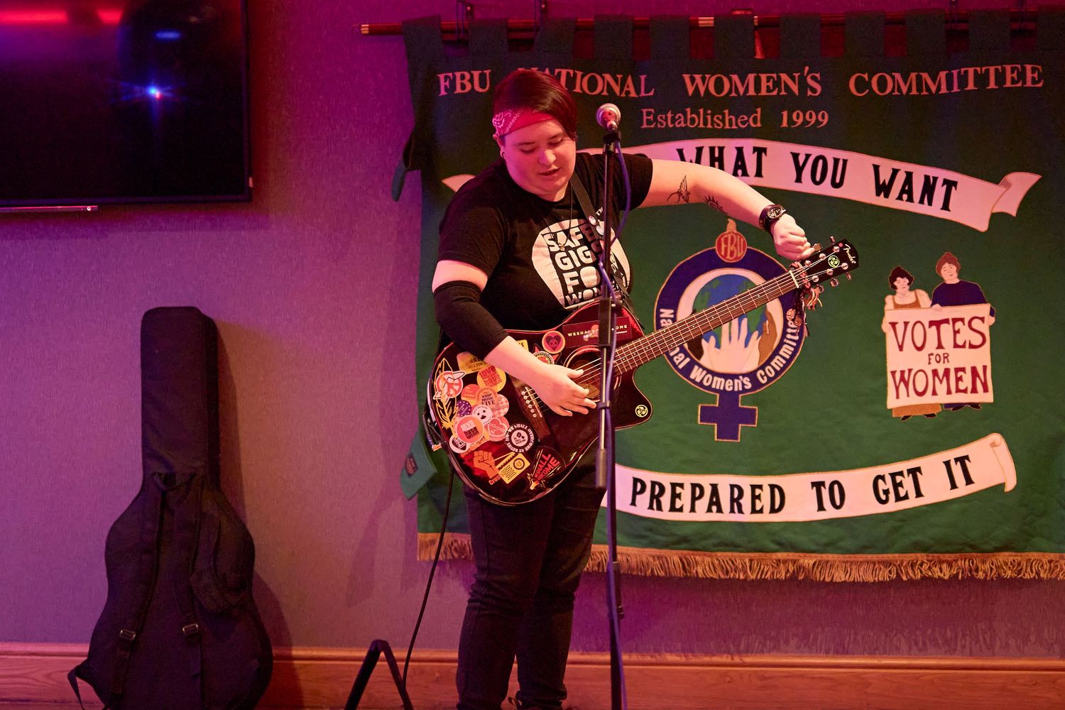 A musician performs on an electric guitar against a backdrop with a suffragette banner in pink and green lighting.