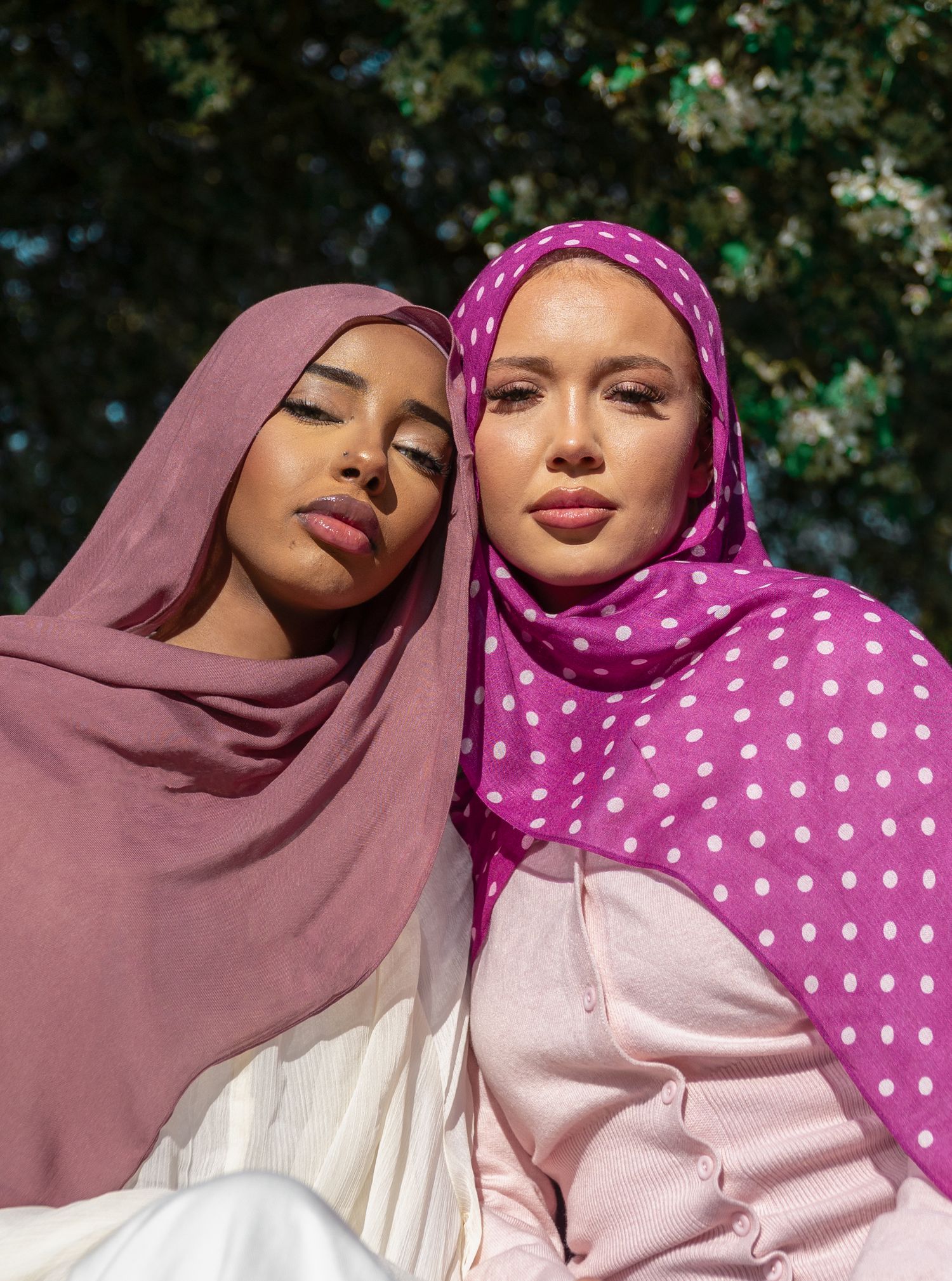 Two women wearing colorful hijabs pose outdoors, one in dusty rose and one in pink polka dot.