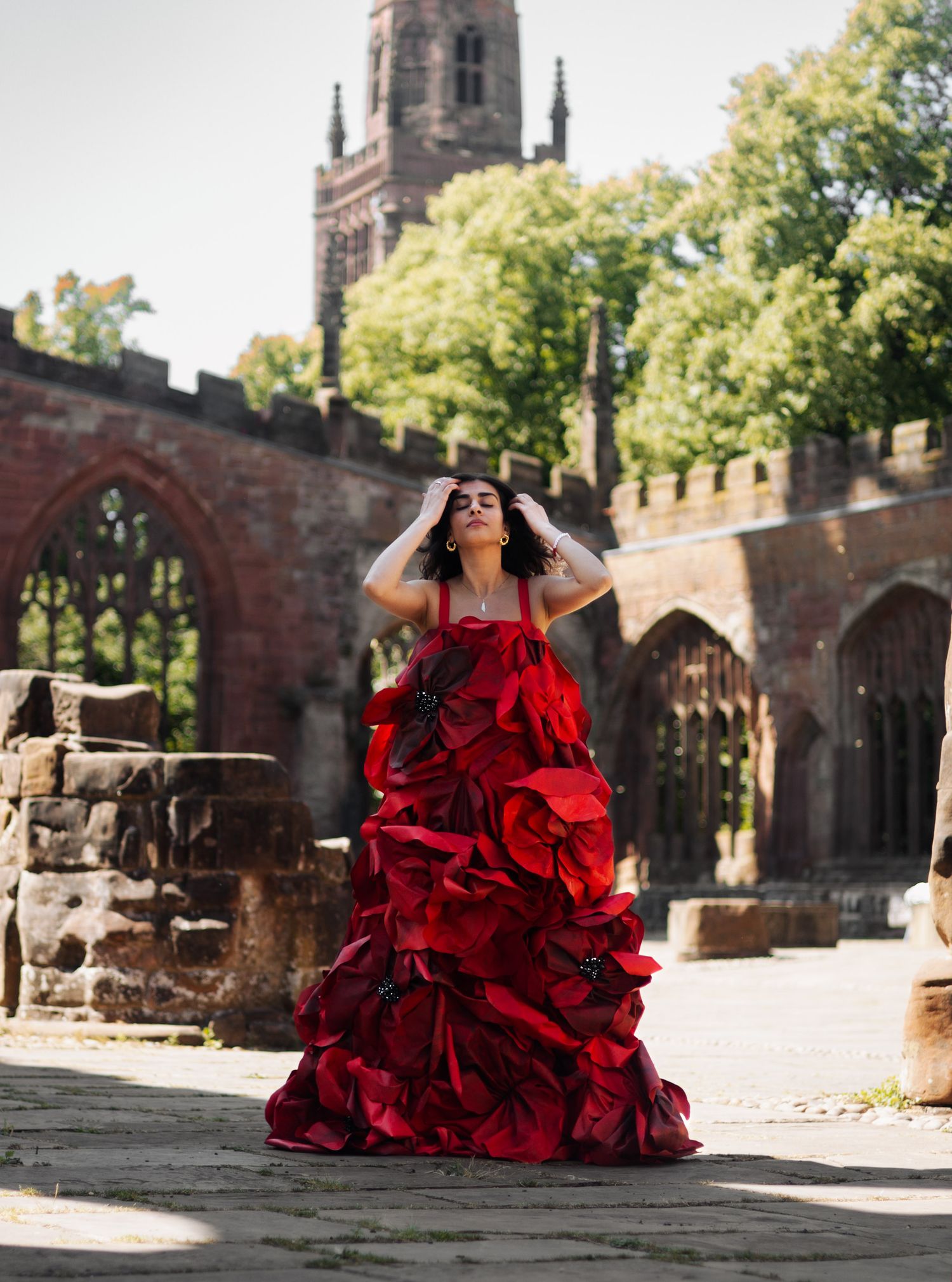 Woman in dramatic red ruffled gown poses at historic stone ruins with Gothic church tower and lush trees in background.