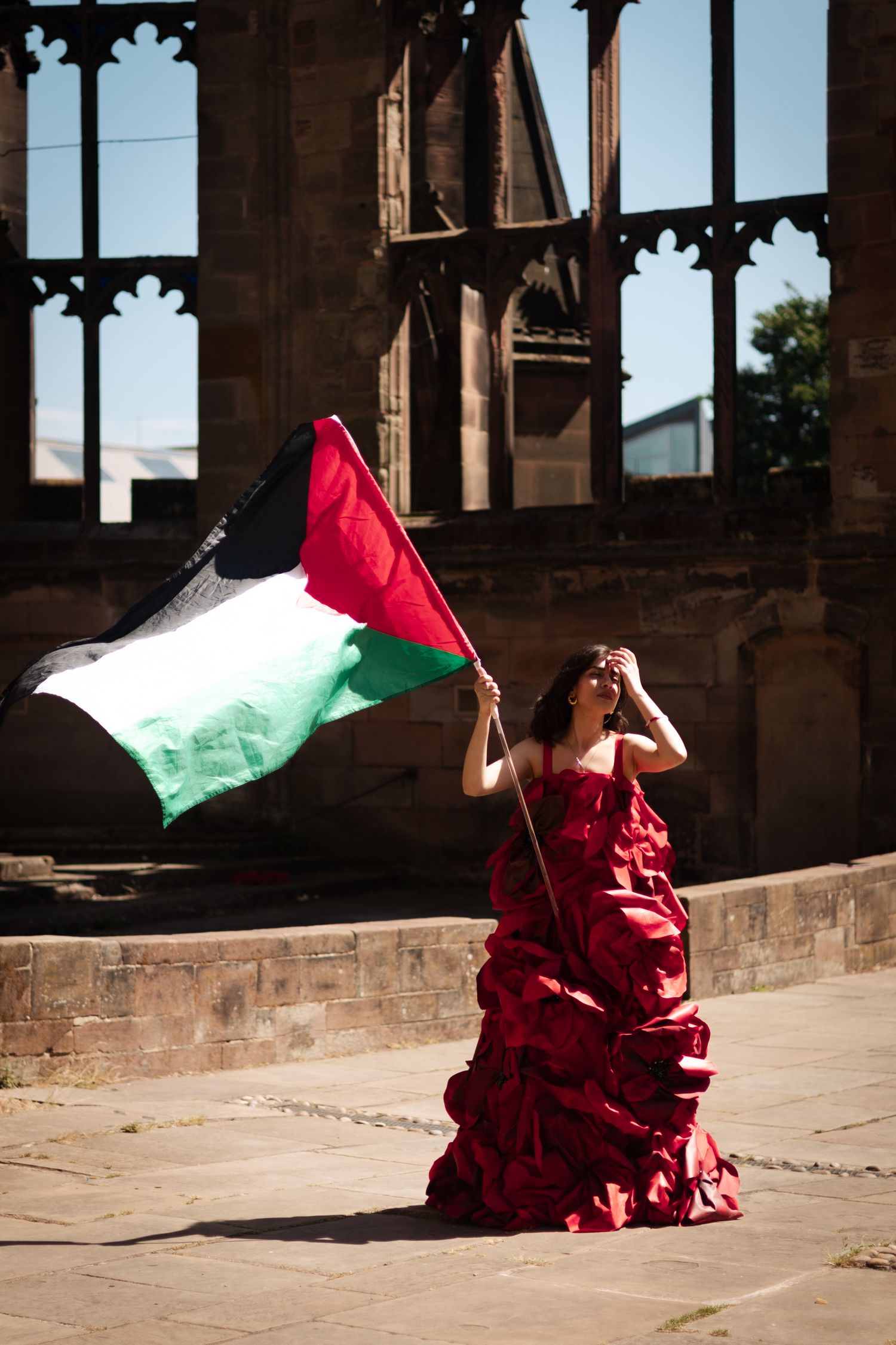 Palestinian flag waves alongside figure in red dress against stone architectural backdrop.