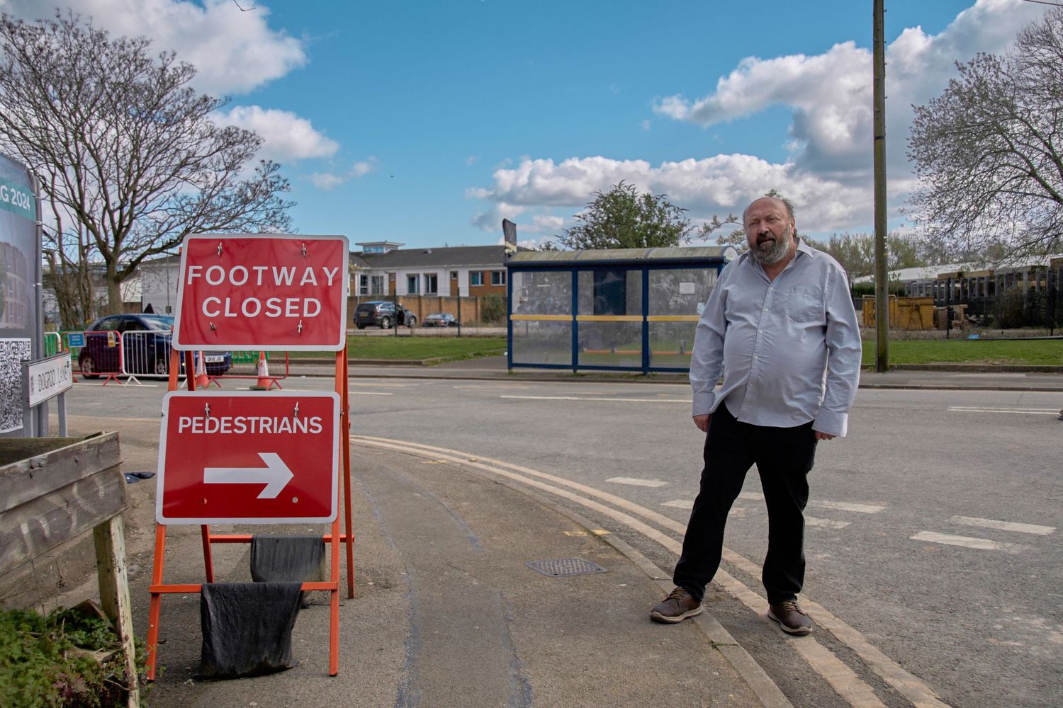 A person stands next to a red 'Footway Closed' sign with pedestrian diversion arrow on a sunny day.