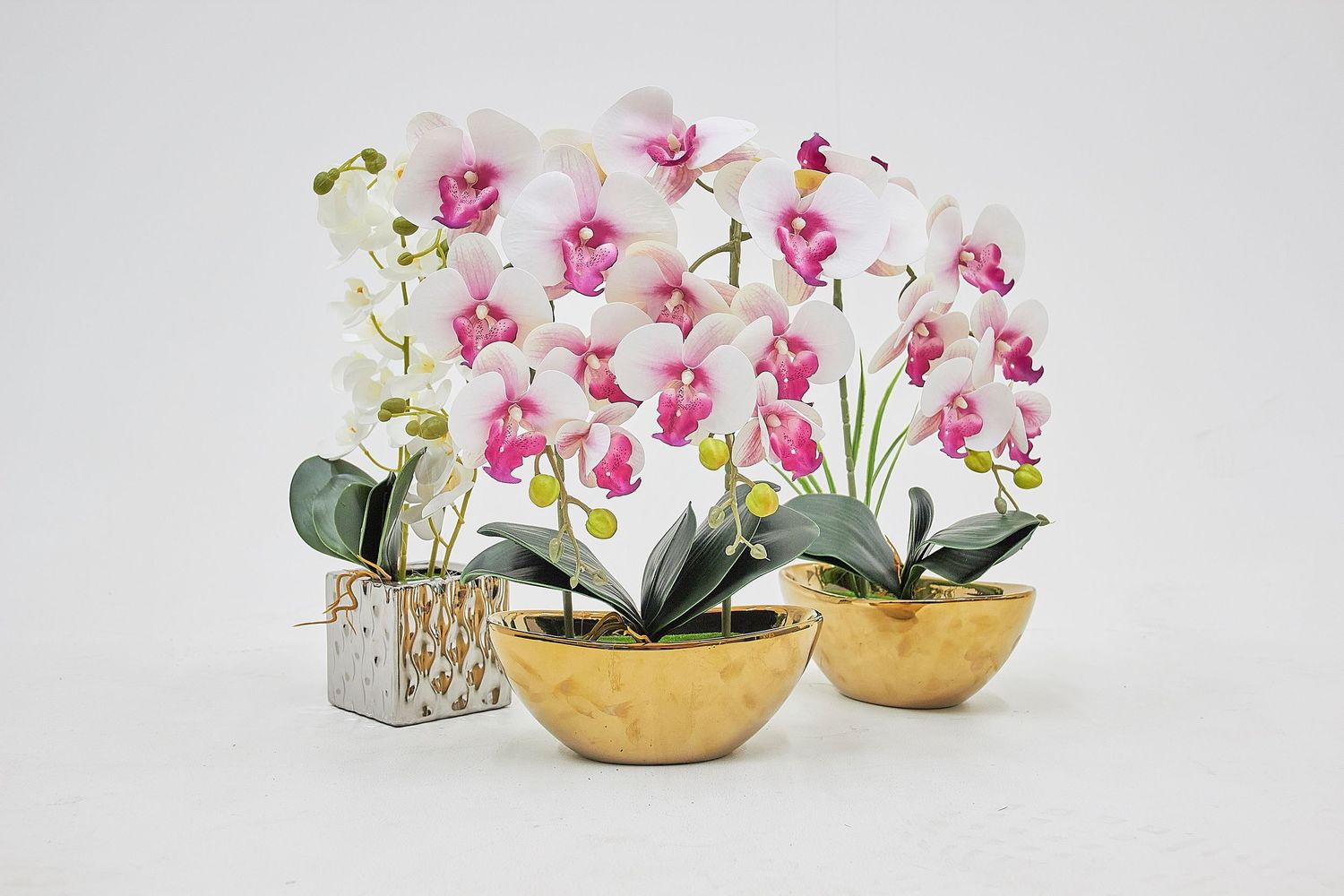Pink orchids and white lilies arranged in gold and metallic patterned decorative containers on a light surface.