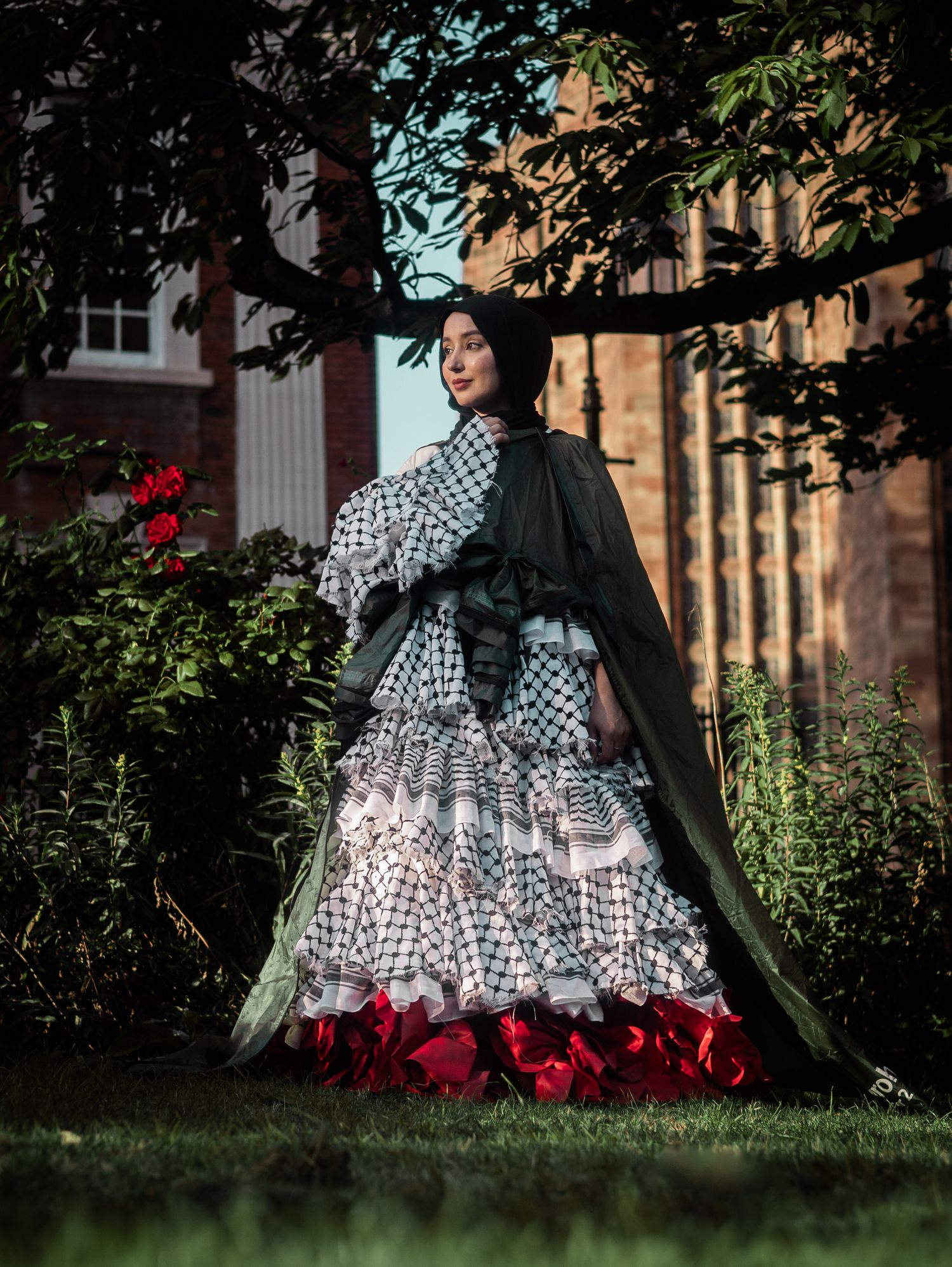 A person in a patterned Victorian-style dress stands in a garden with red flowers and stone buildings in the background.