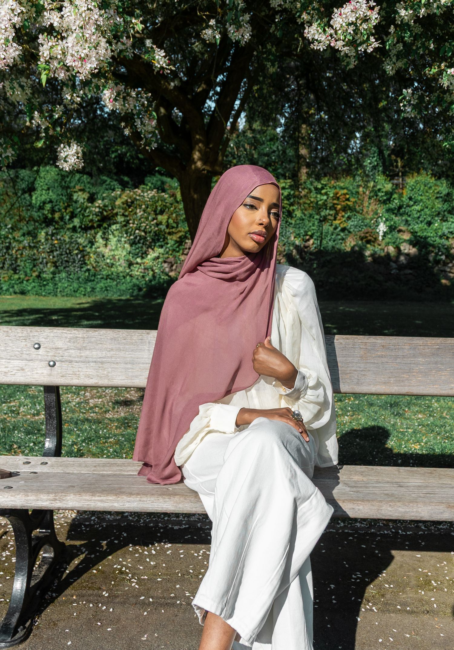 Woman wearing a mauve hijab and white outfit sitting gracefully on a park bench surrounded by blooming trees.