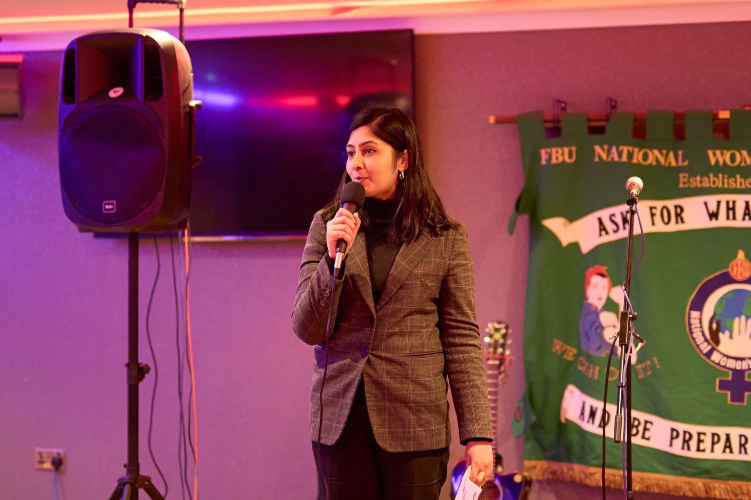 A performer in a brown blazer speaks into a microphone on stage with pink and purple lighting in the background.