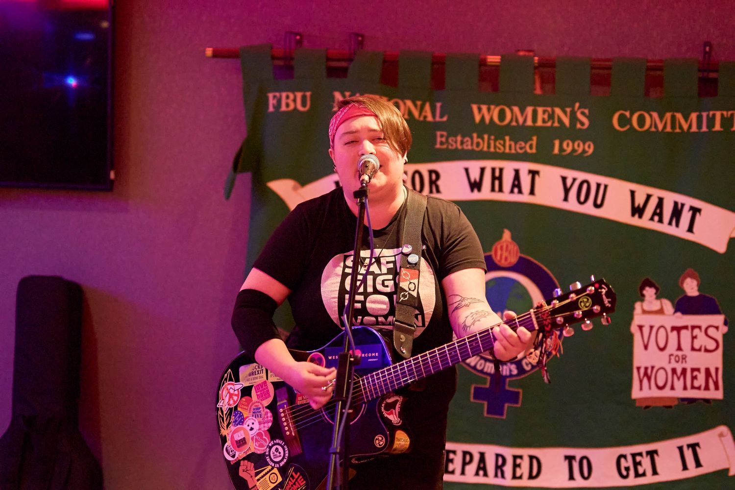 A person performs with a guitar on stage with a feminist political banner visible in the background during a show.