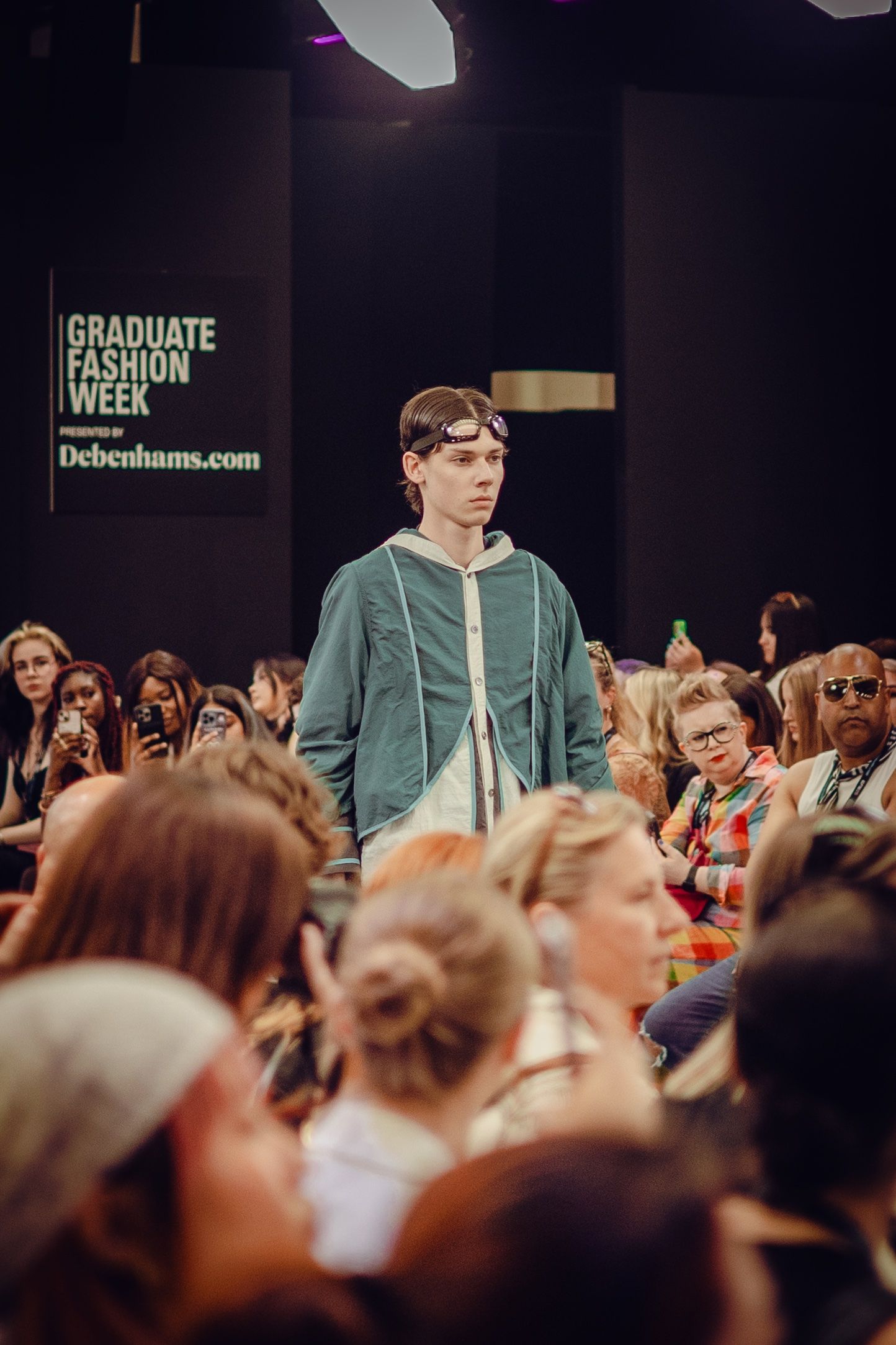 Model in teal jacket walks runway at Graduate Fashion Week event.