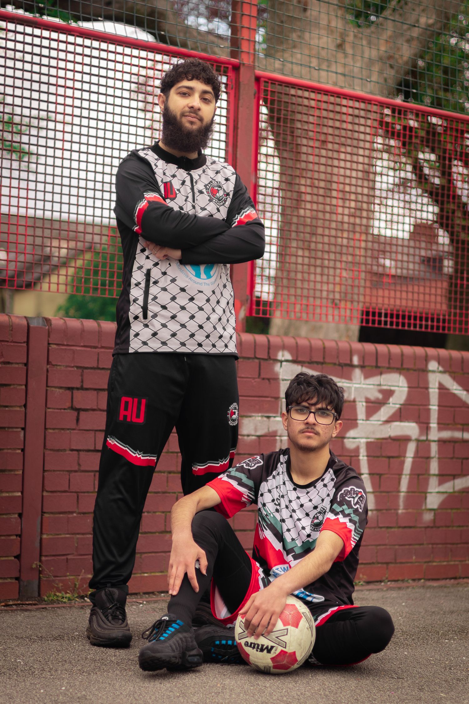 Two players in matching black and white uniforms pose with a soccer ball against a red fence.