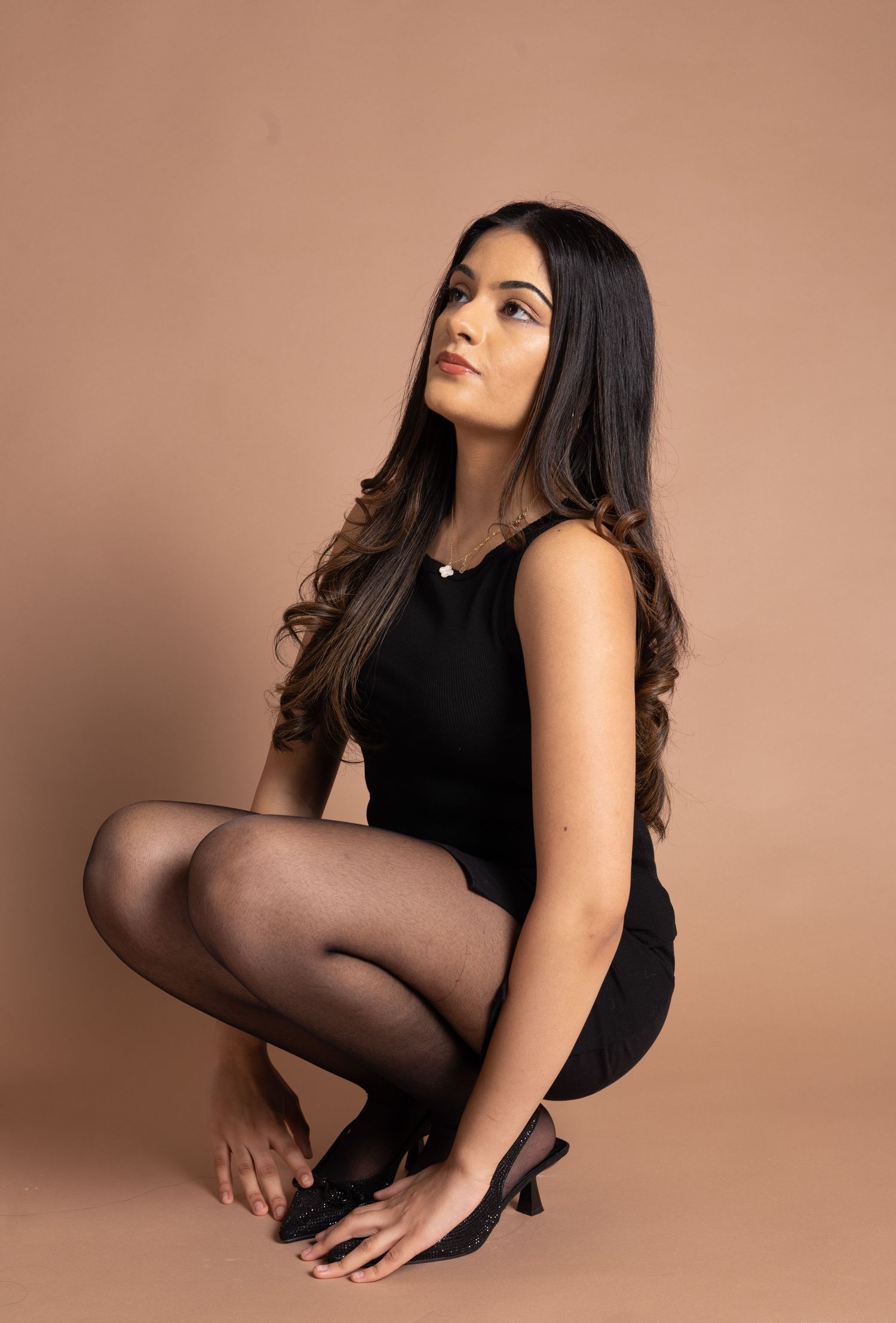 Young woman in black dress and tights sitting gracefully against a beige studio background.