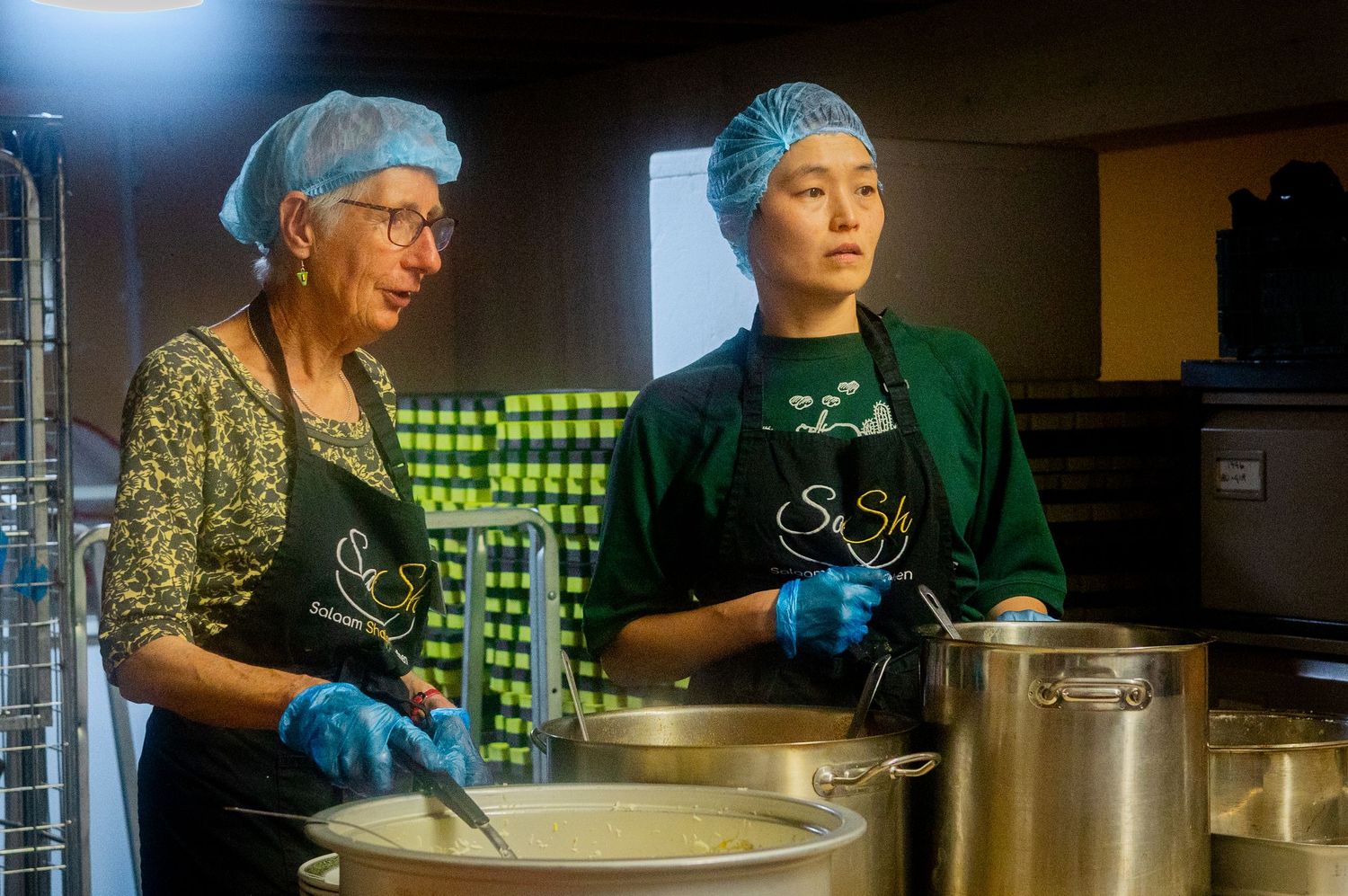 Two volunteers in hairnets and aprons work together ladling soup in a community kitchen.