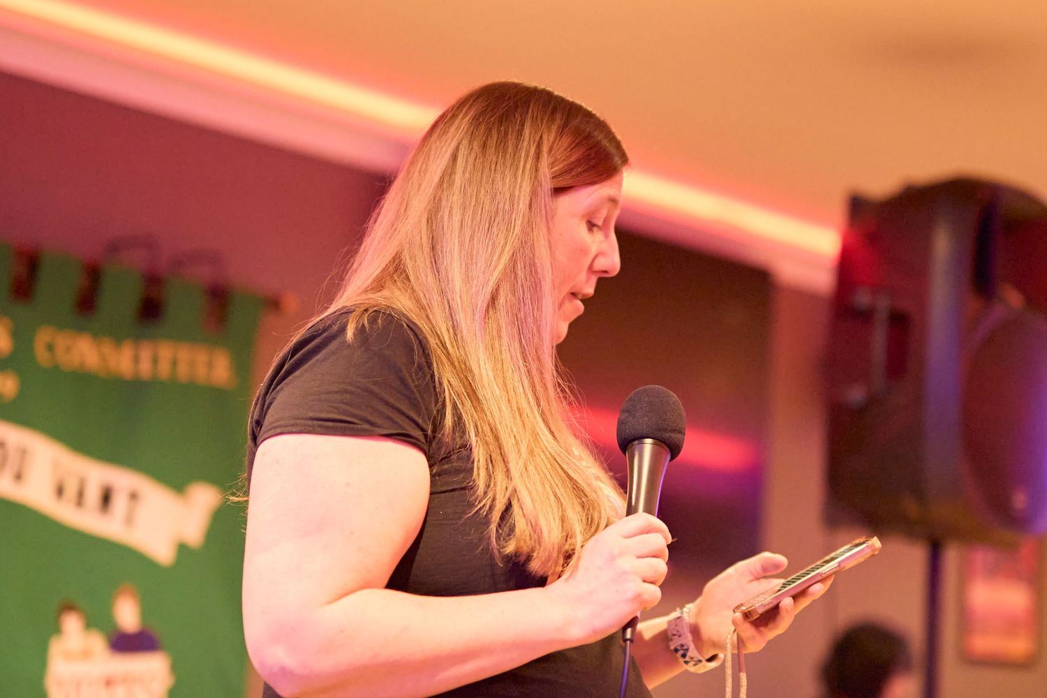 A person with long blonde hair speaks into a microphone while reading from a paper at an event.