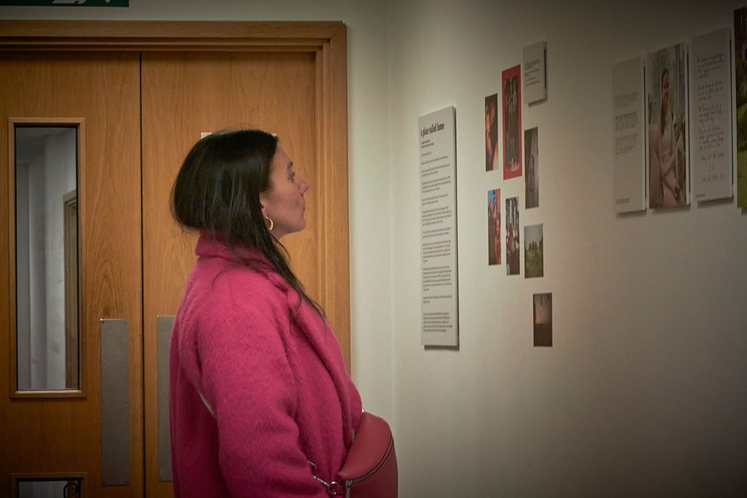 A person in a pink sweater walks down a dimly lit hallway past doors and wall postings.