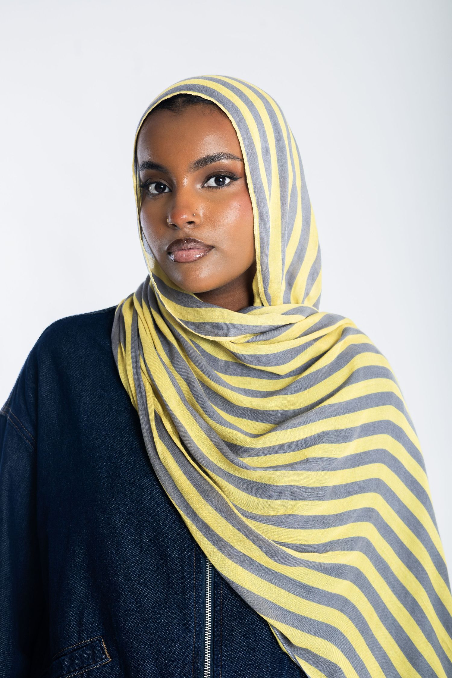 Young woman wearing a yellow and gray striped hijab with a denim jacket against a white background.