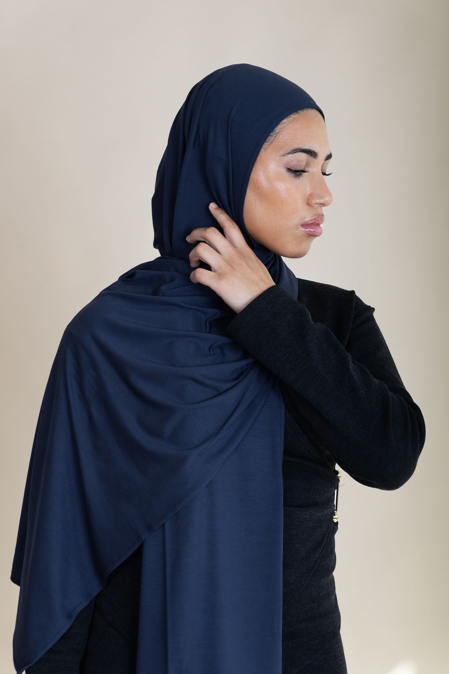 Side profile portrait wearing a navy blue hijab and black sweater against a neutral beige background.
