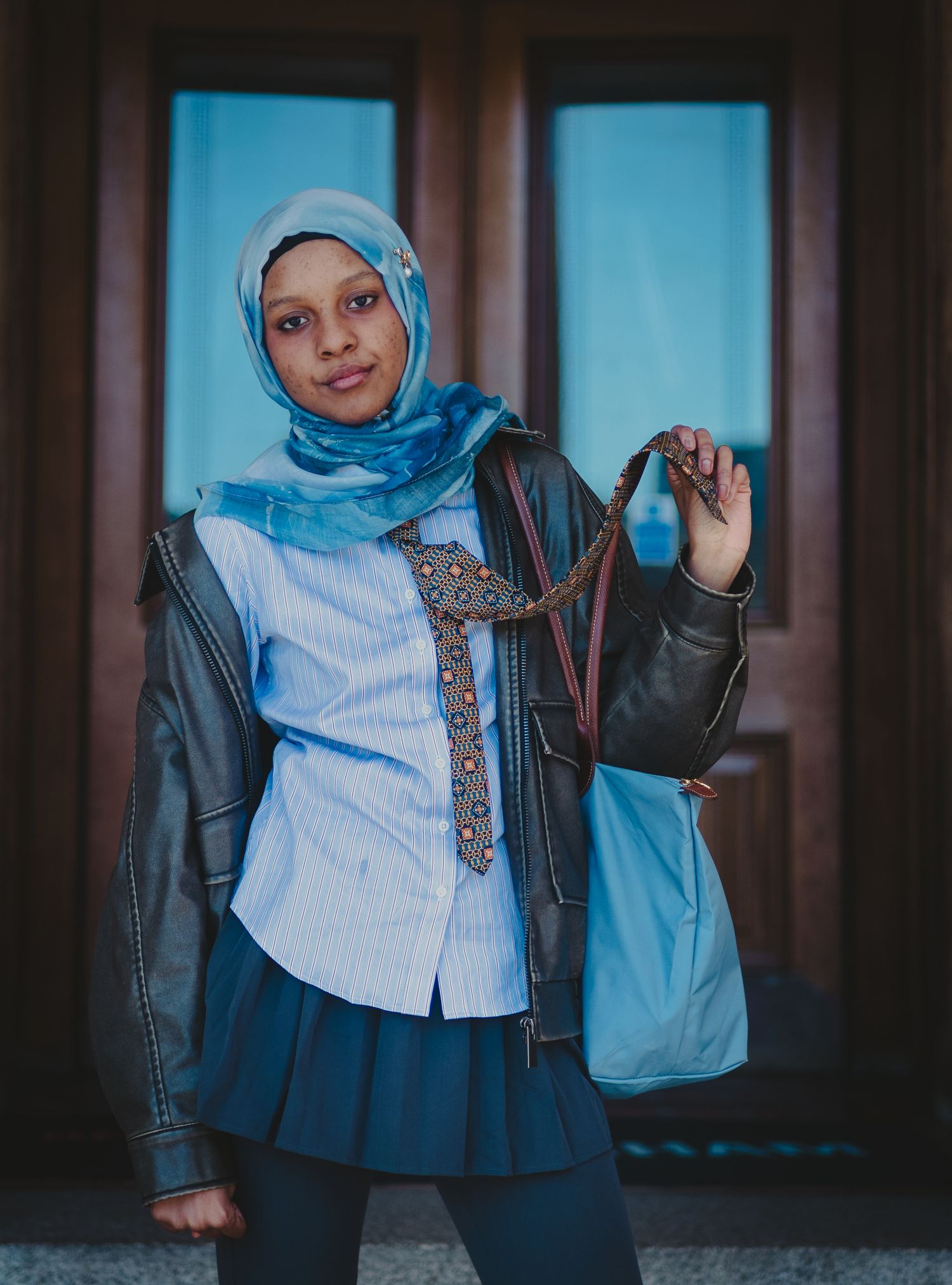 Stylish outfit featuring a leather jacket, blue hijab, button-down shirt and patterned tie against wooden doors.