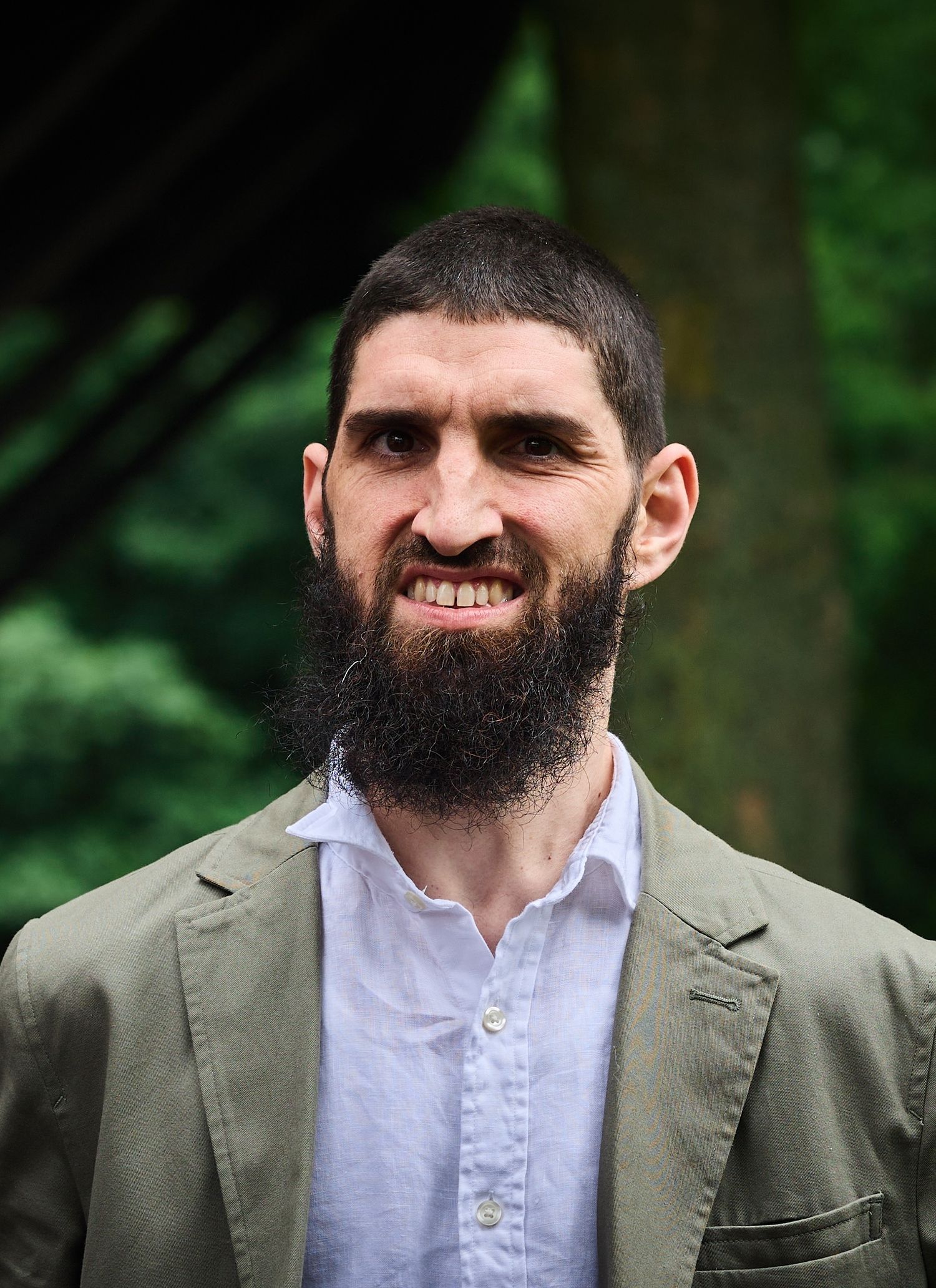Bearded man in olive blazer smiling outdoors with green trees in background.