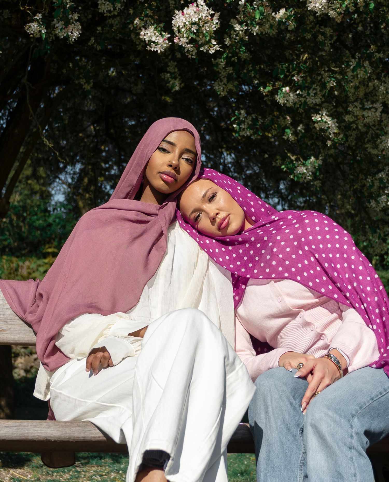 Two women wearing colorful hijabs sit together on a bench outdoors beneath a flowering tree.
