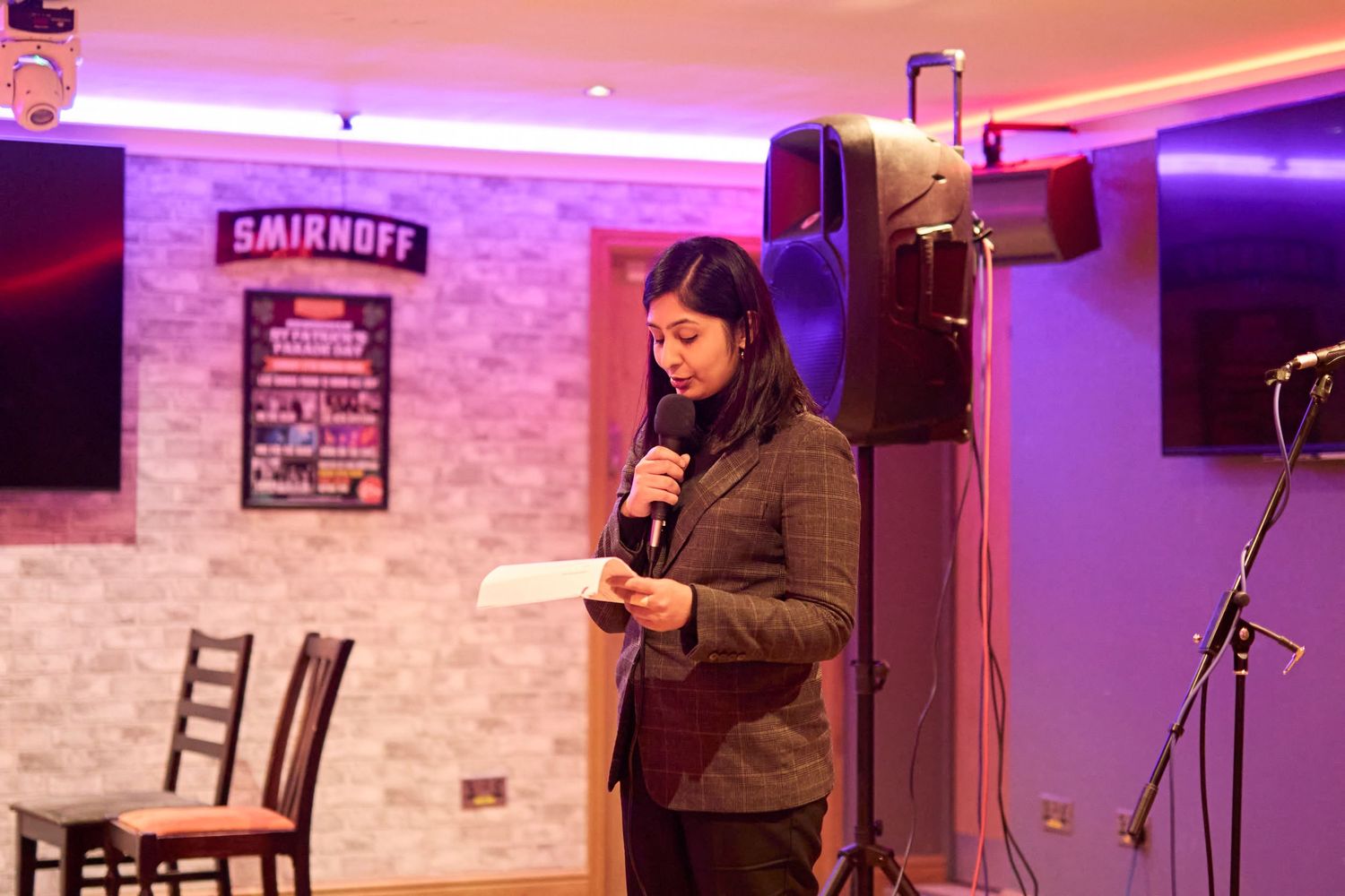 A performer speaks into a microphone at a comedy club with purple lighting and exposed brick walls.