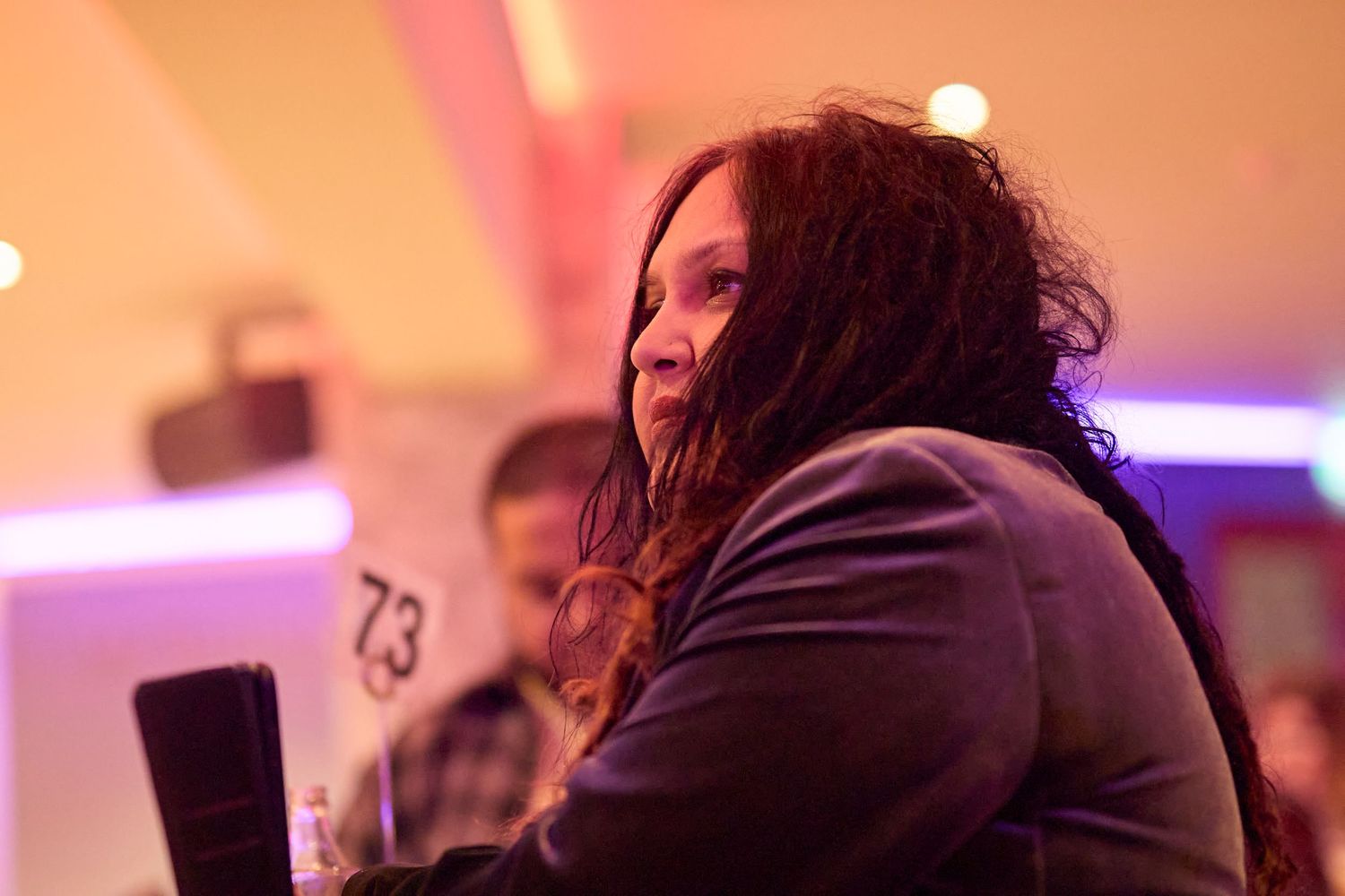 A person with long dark hair sits in profile view under neon lighting at a dimly lit venue.