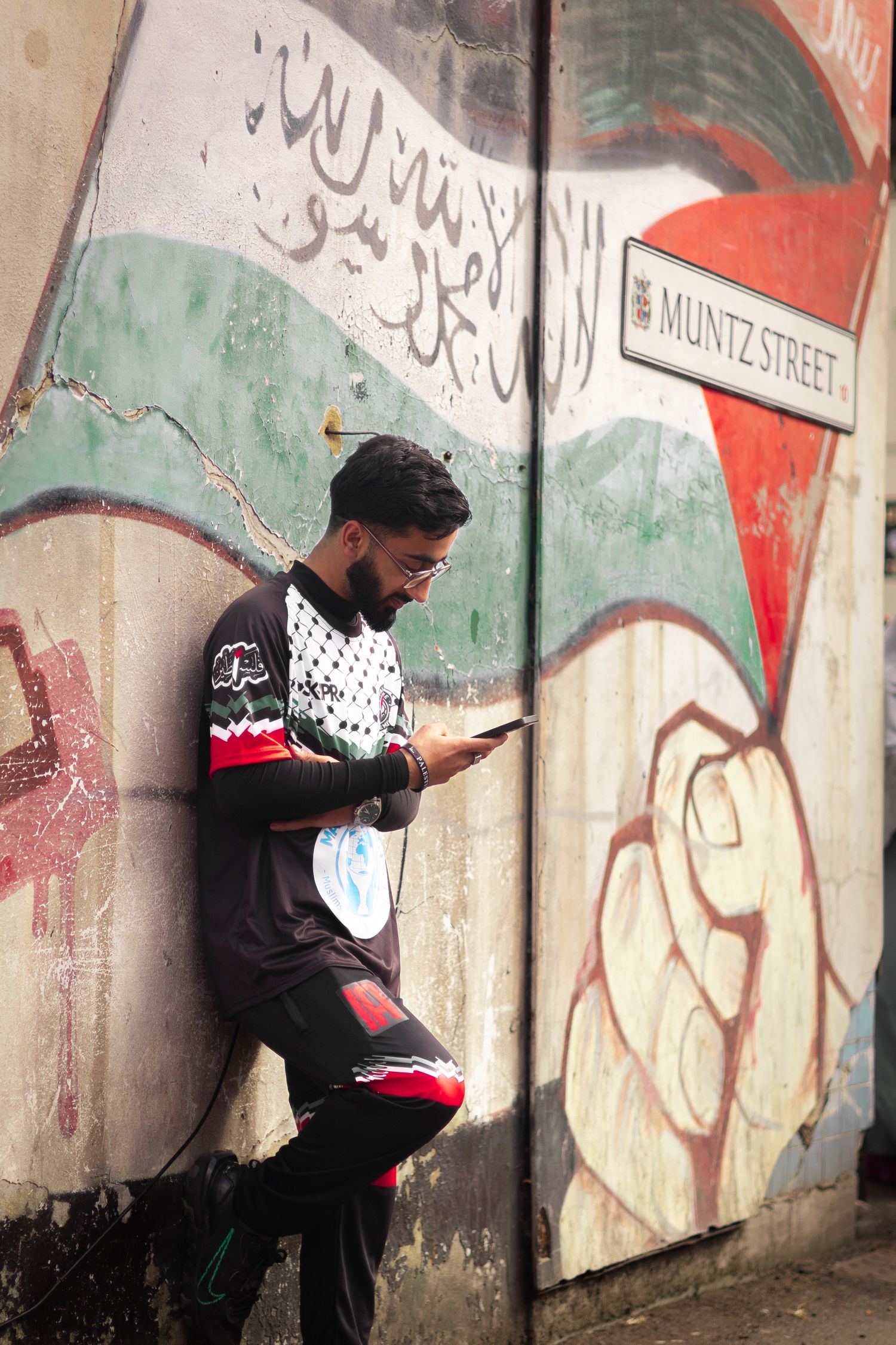 Man in black jersey leaning against a Palestinian mural on Muntz Street, looking at his phone.