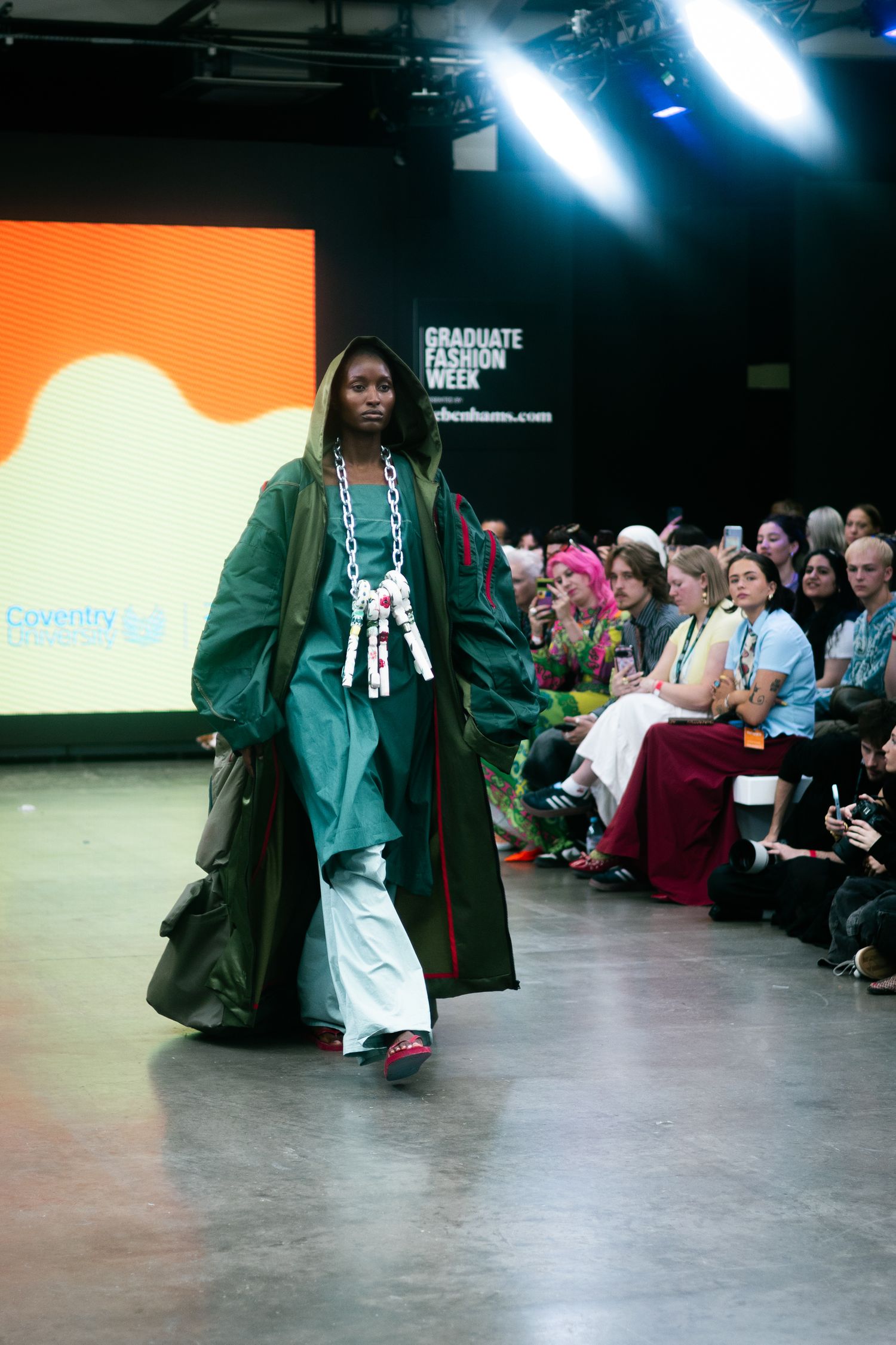 Model walks runway in green oversized coat with white decorative details against orange backdrop.