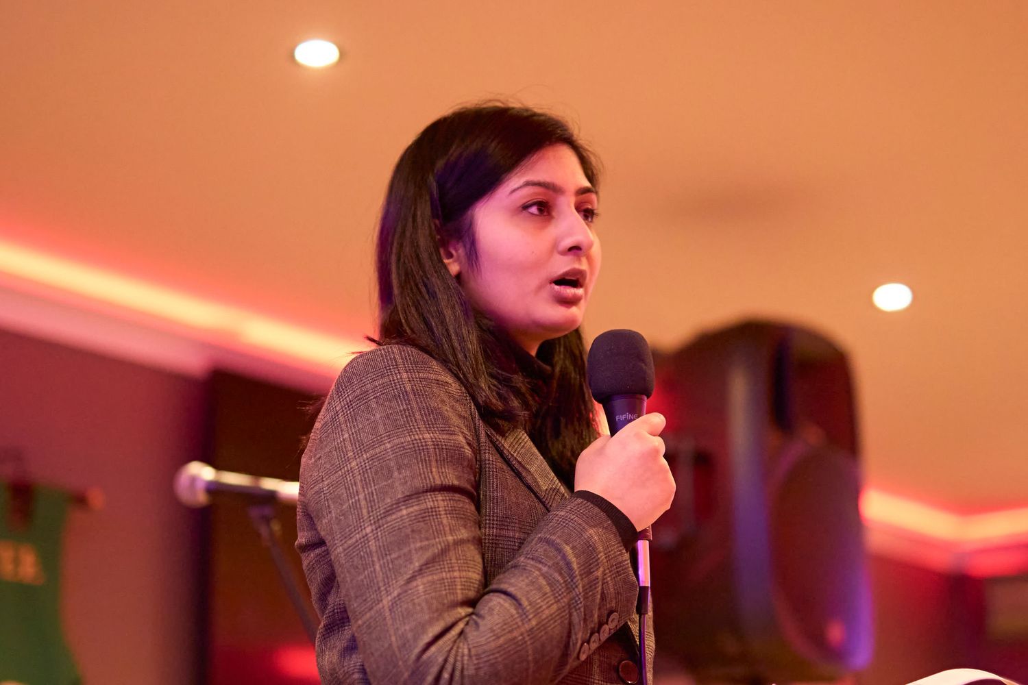 Someone speaking into microphone during performance in room with red ambient lighting and plaid blazer.