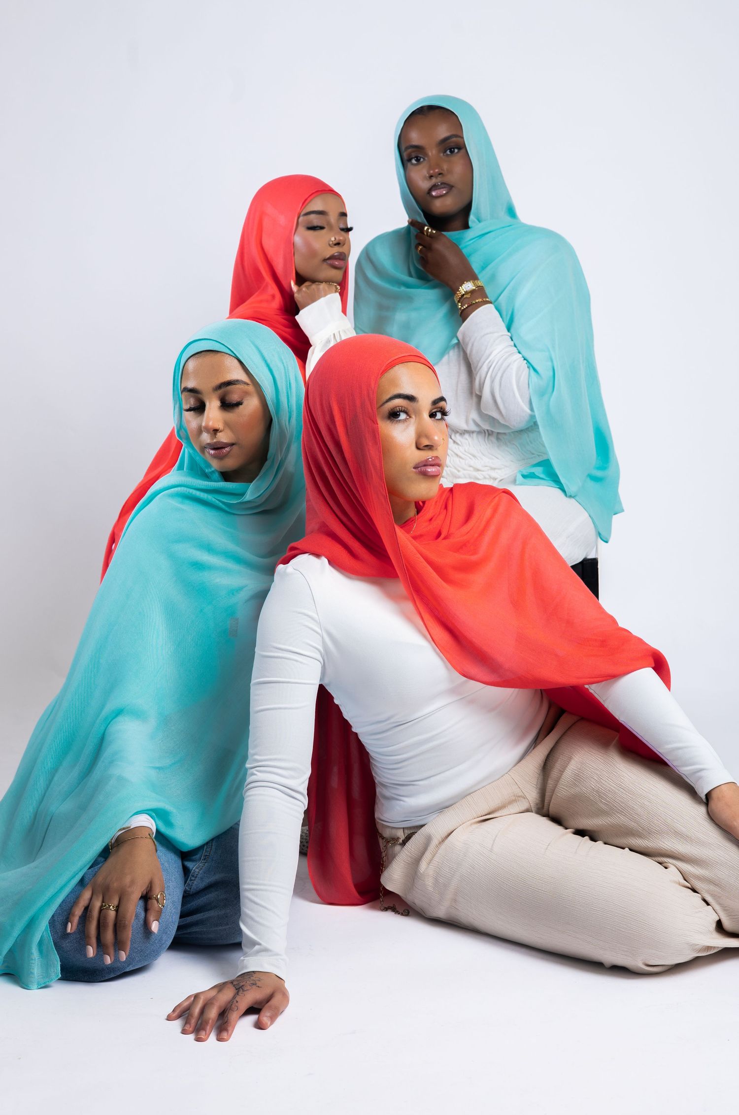 Four women wearing colorful teal and coral hijabs pose together in a studio setting against a white background.