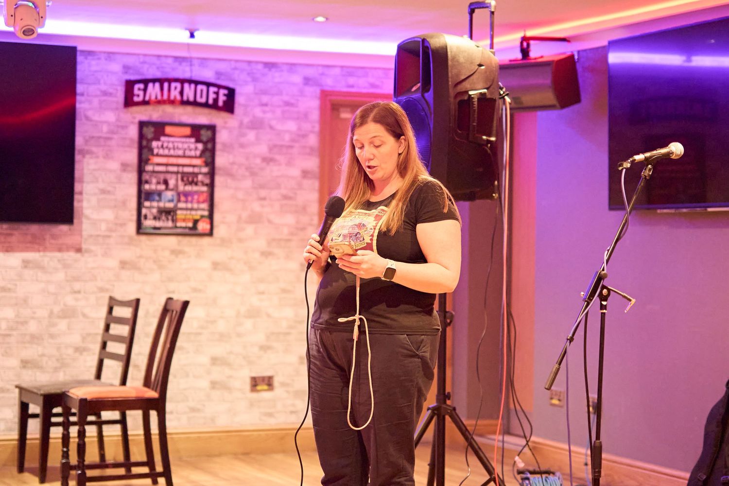 A performer stands on stage with a microphone in a dimly lit venue with purple lighting and exposed brick walls.