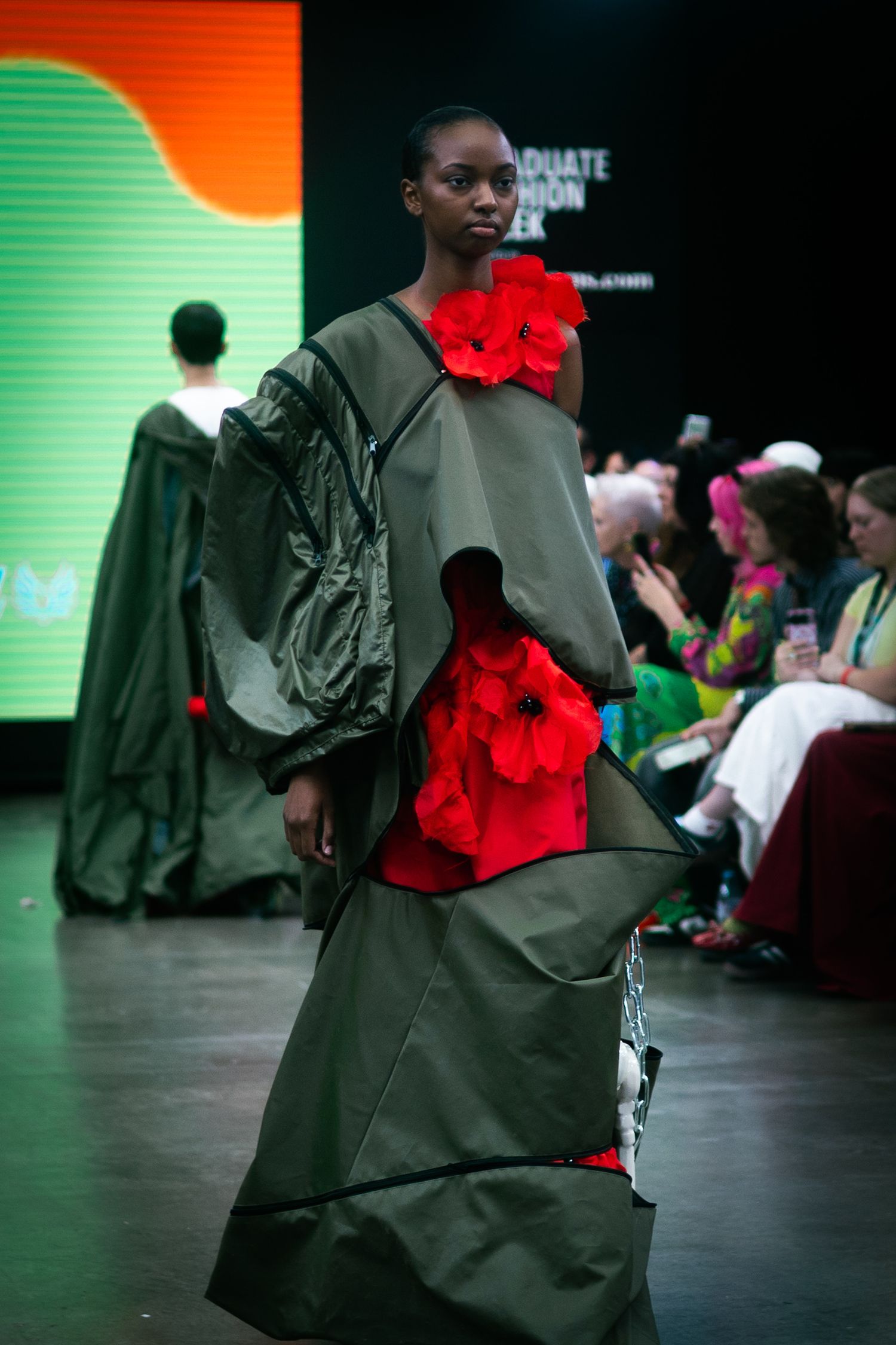 Model showcases olive green gown with dramatic red floral accents on fashion runway.