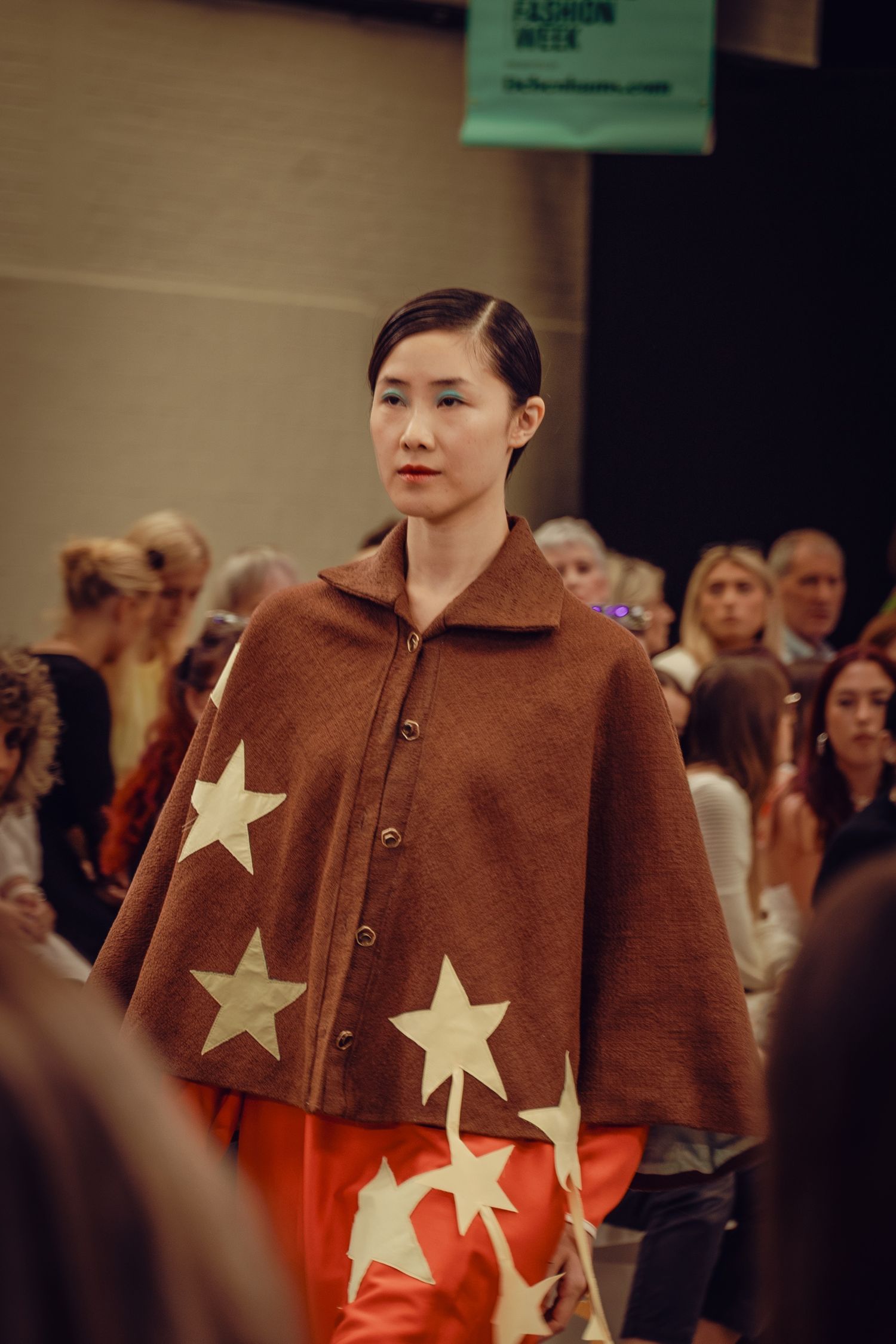 Model showcases brown cape with star designs and orange pants on fashion runway.