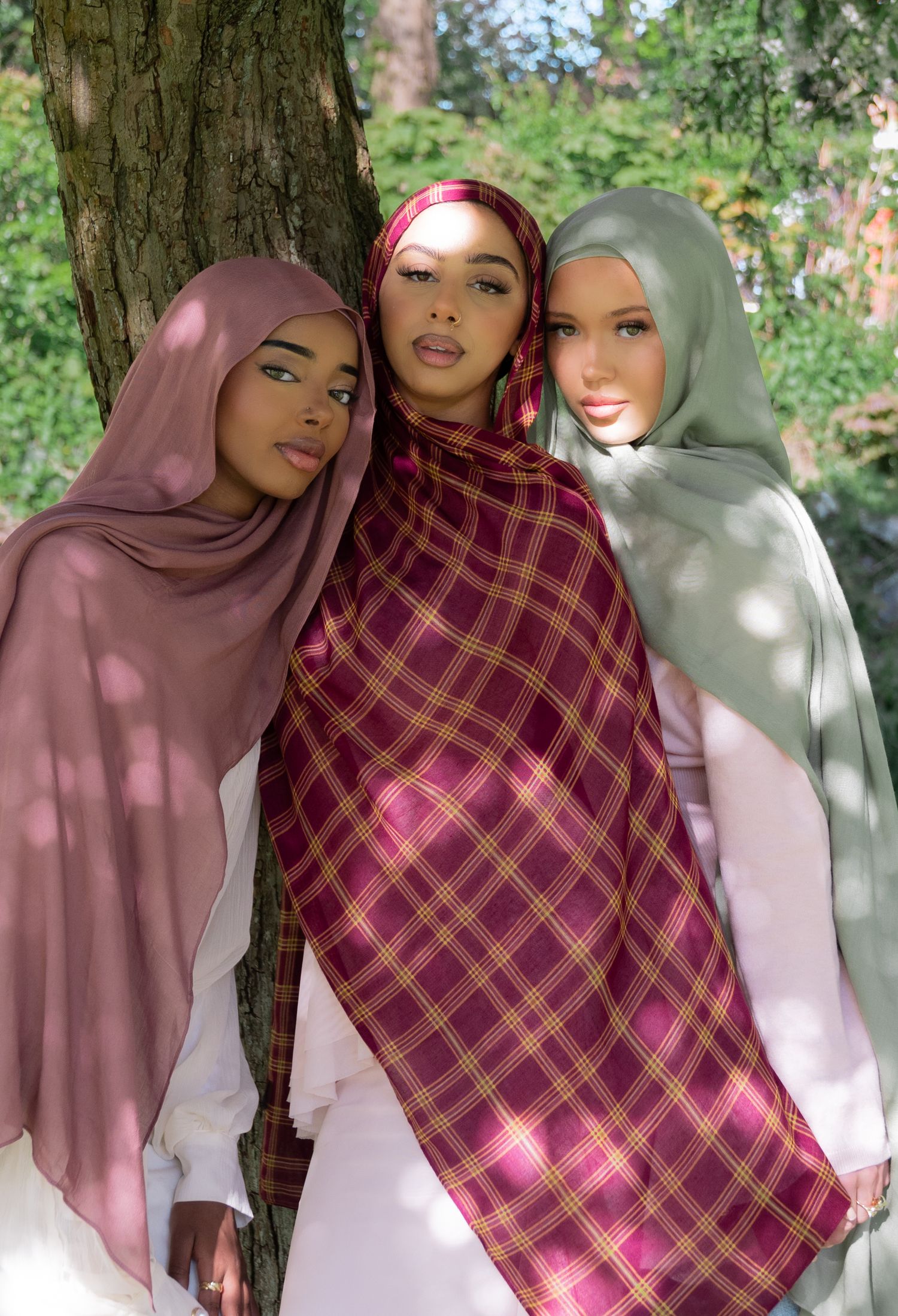 Three women wearing colorful hijabs pose together outdoors near a tree, showcasing pink, plaid, and sage green styles.