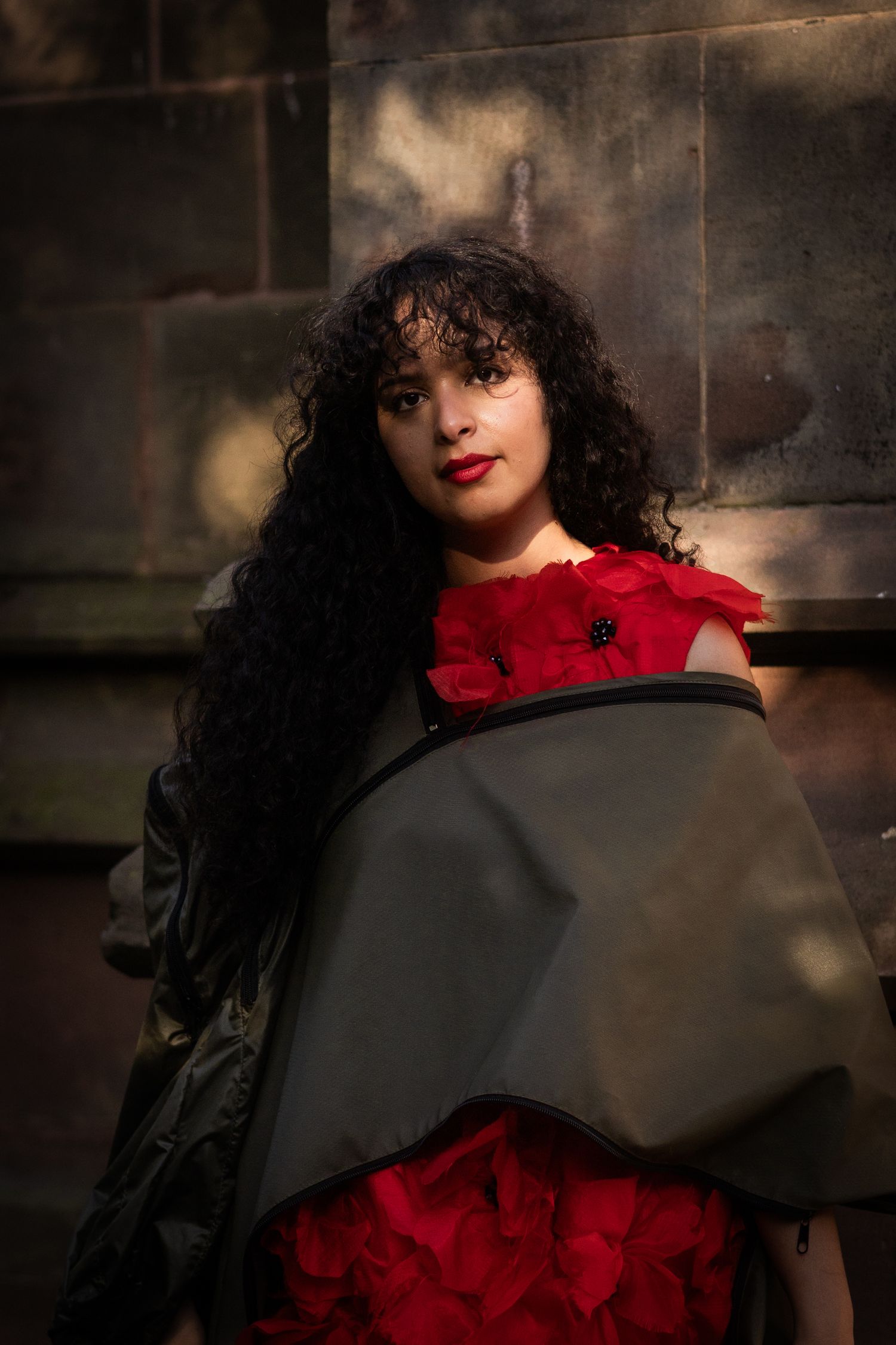 Woman with curly black hair in red floral dress and dark cape poses against stone wall.