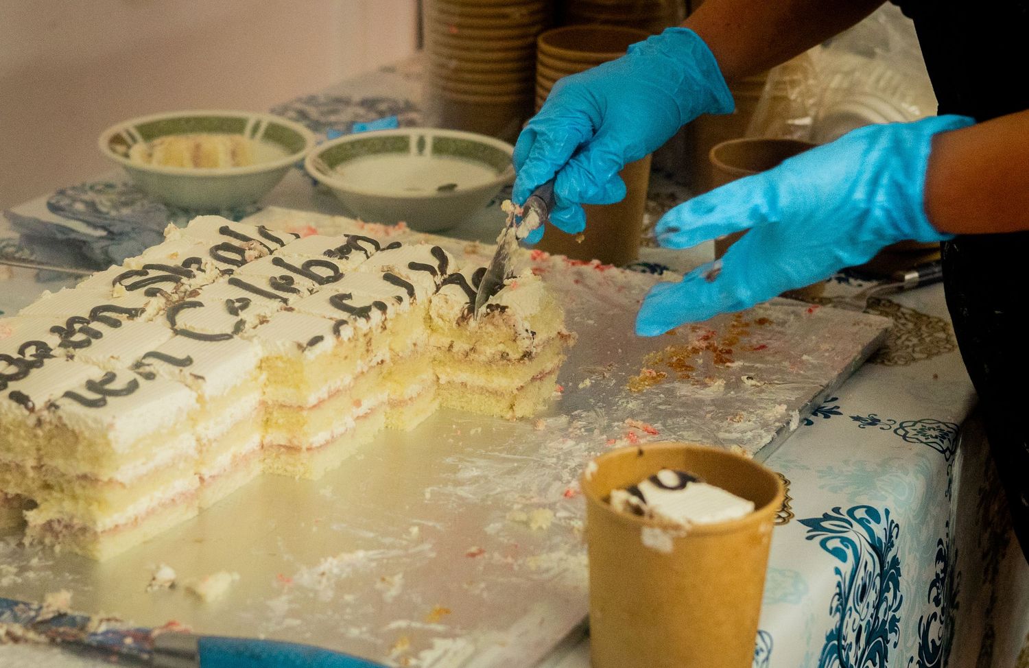 Hands wearing blue gloves slice into a layered cream cake on a decorated table.