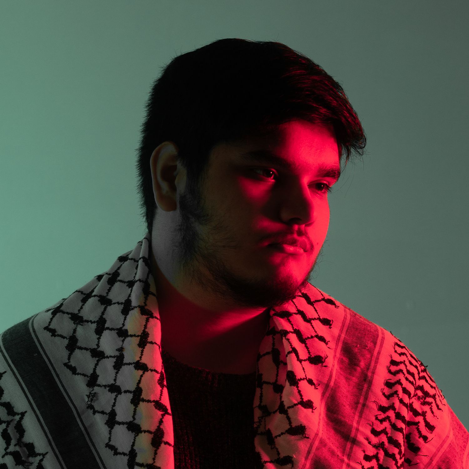 Dramatic portrait with red lighting showing traditional keffiyeh pattern in shadow.