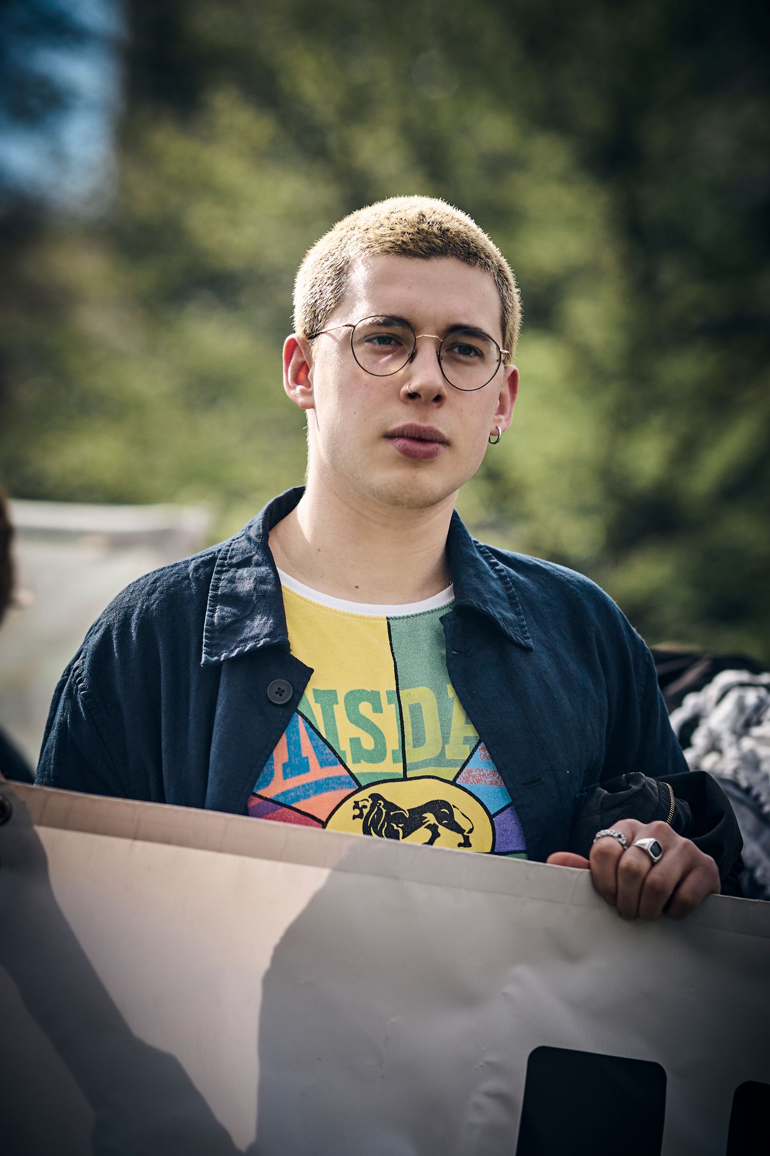 Young protestor in denim jacket and glasses holds banner at outdoor demonstration.