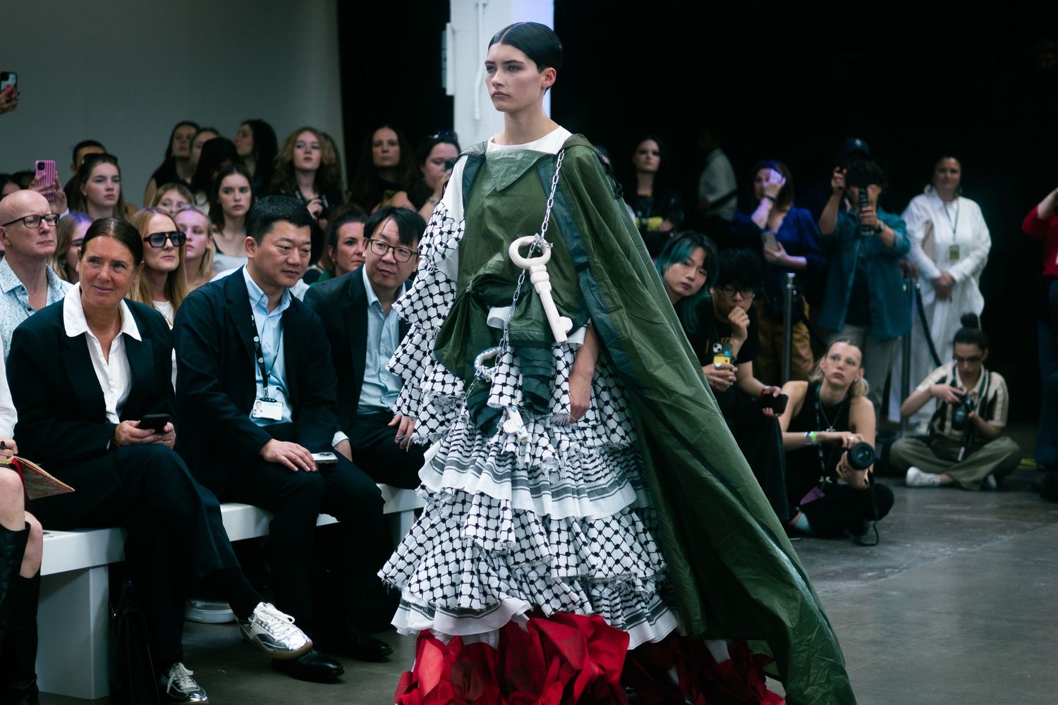 Fashion model wearing polka dot ruffled dress with green cape on runway surrounded by audience.
