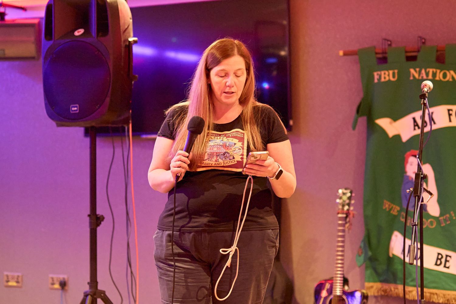 A performer in black overalls stands at a microphone on stage with purple lighting and musical equipment visible.