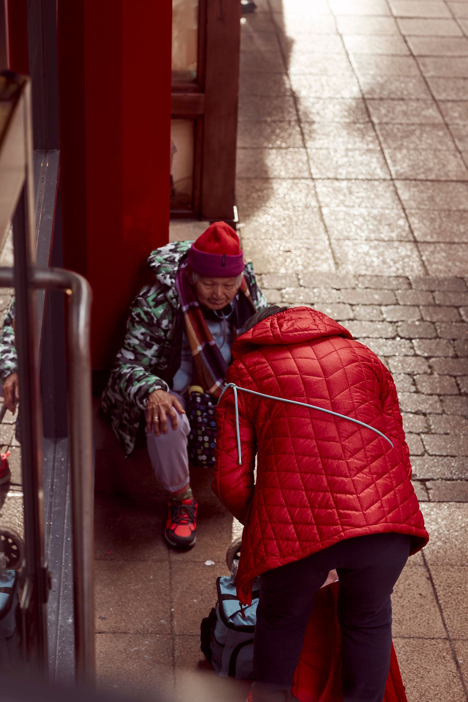 Two people interact on a tiled street, one wearing a bright red quilted jacket.