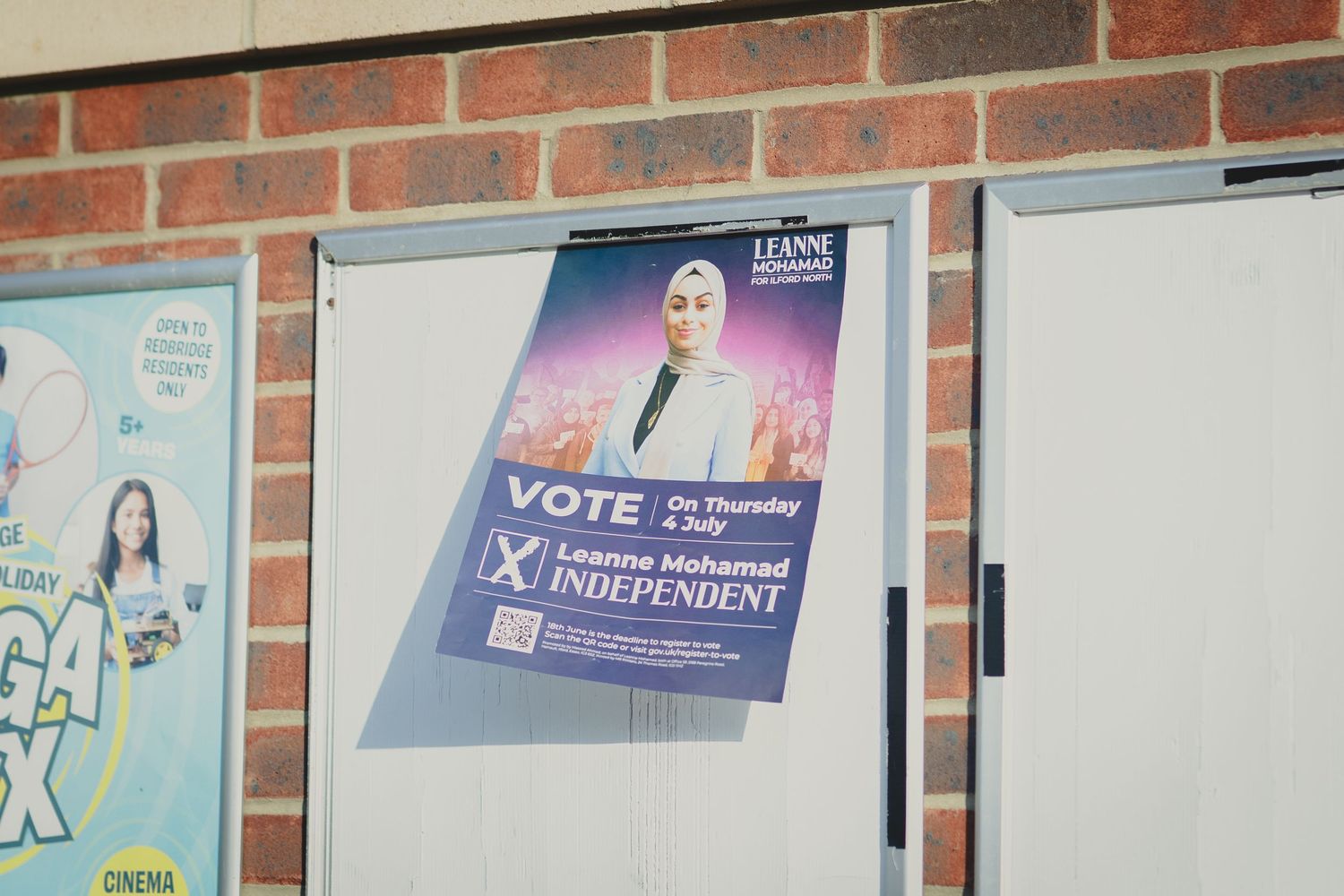 A campaign poster hangs on a brick wall with other posters visible in the background.