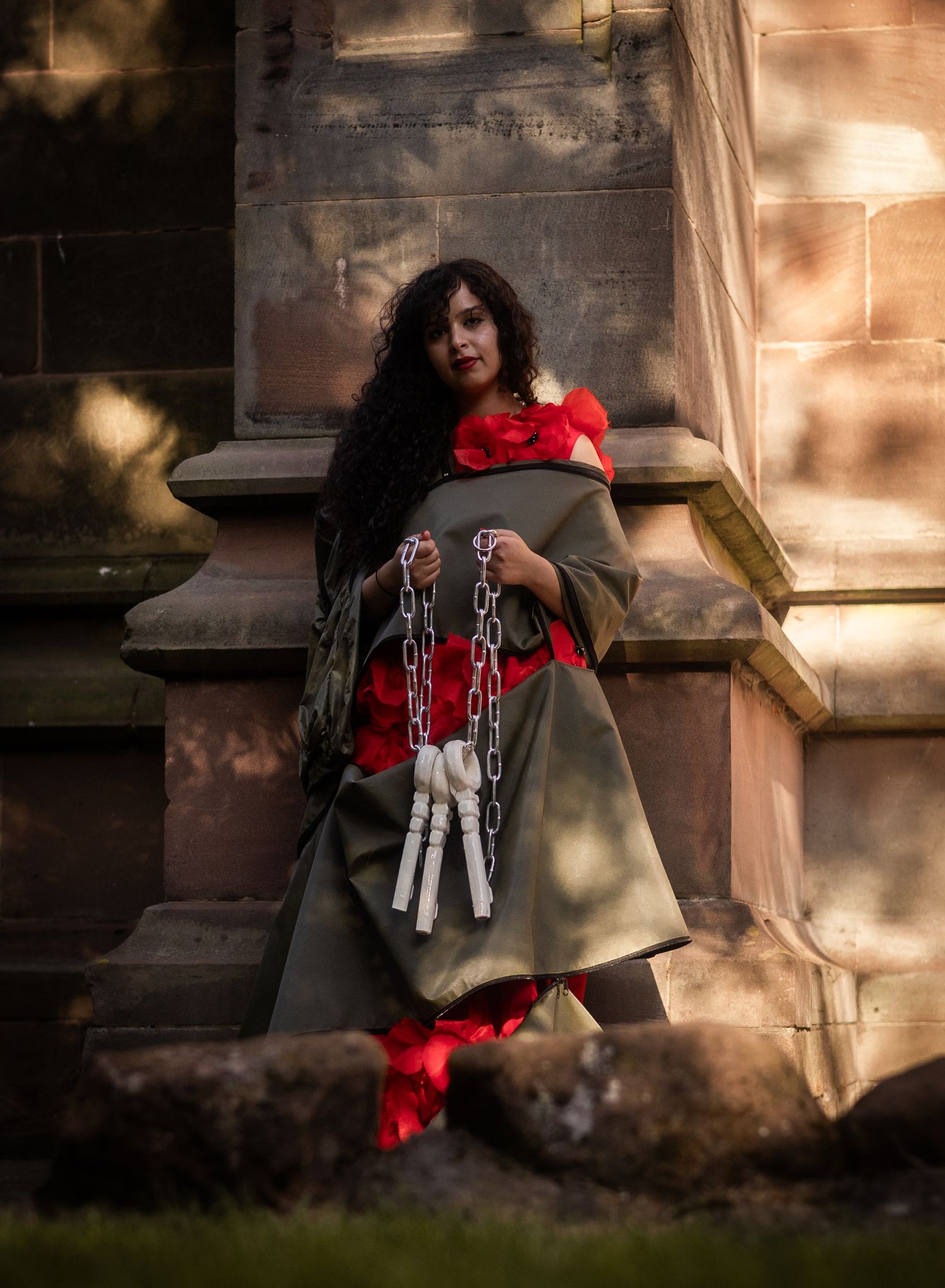 Figure wearing dark clothing and red accents with chains stands dramatically against stone wall in moody lighting.
