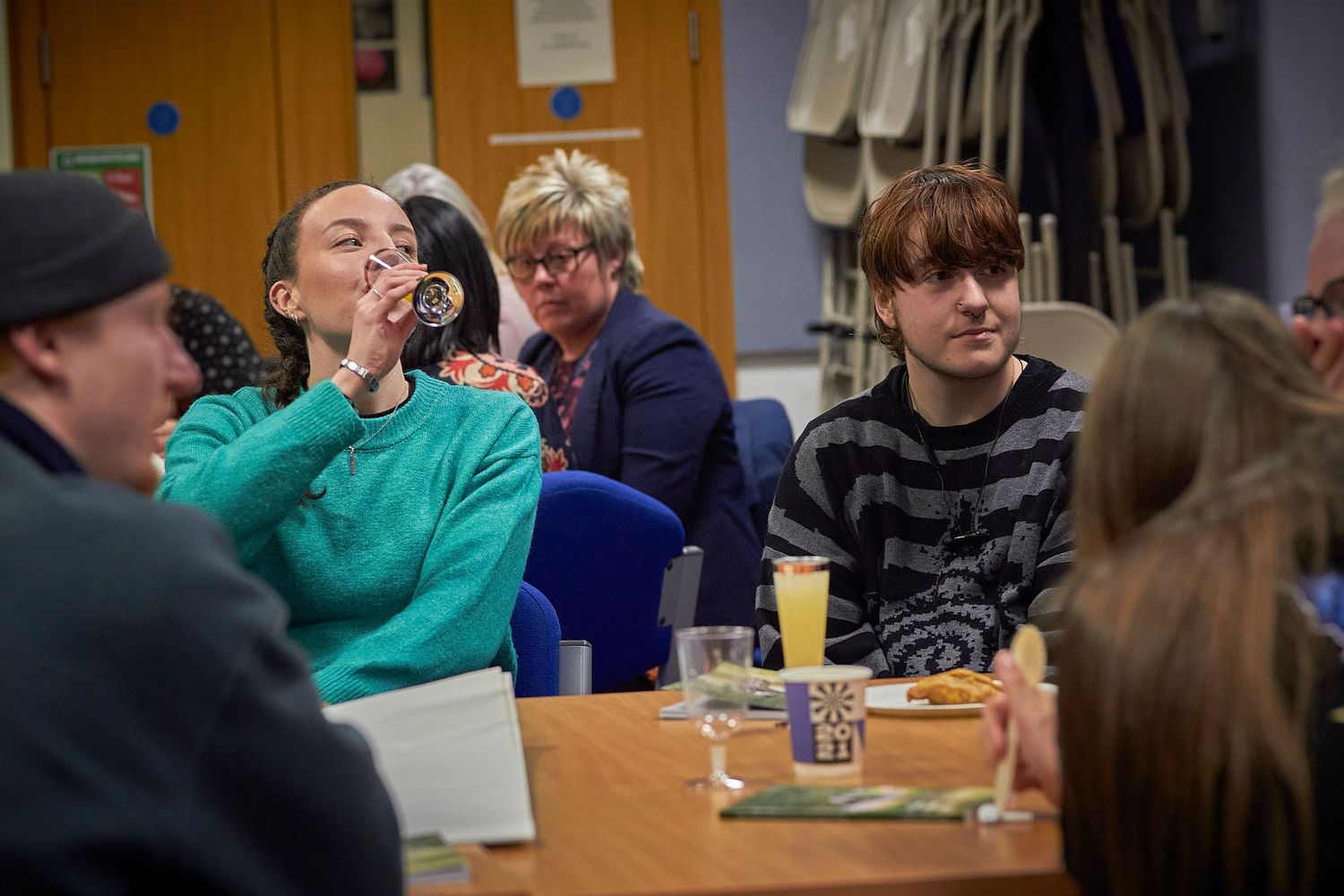 People gathered around a table in a casual indoor setting with one person wearing a teal sweatshirt.