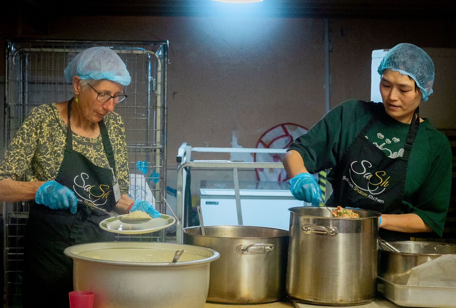 Kitchen volunteers prepare food in large pots under bright overhead lighting.
